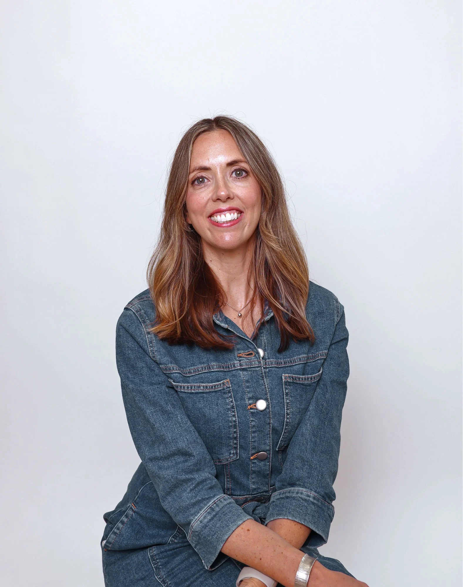 A woman with long wavy brown hair wearing a dark denim jacket, smiling in front of a plain white background.