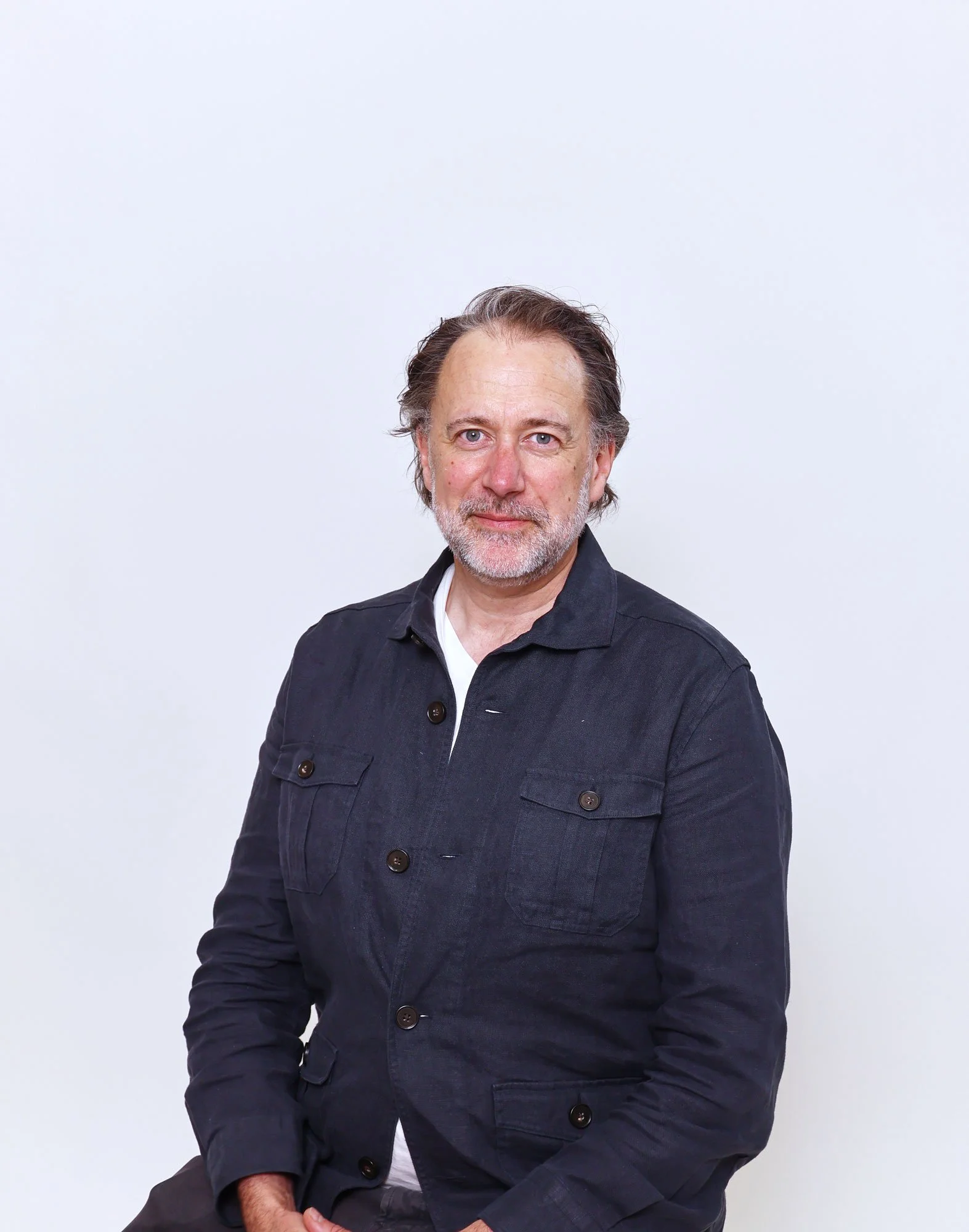 A middle-aged man with gray hair, blue eyes, and a beard, wearing a dark denim jacket and a white shirt, standing against a plain white background.
