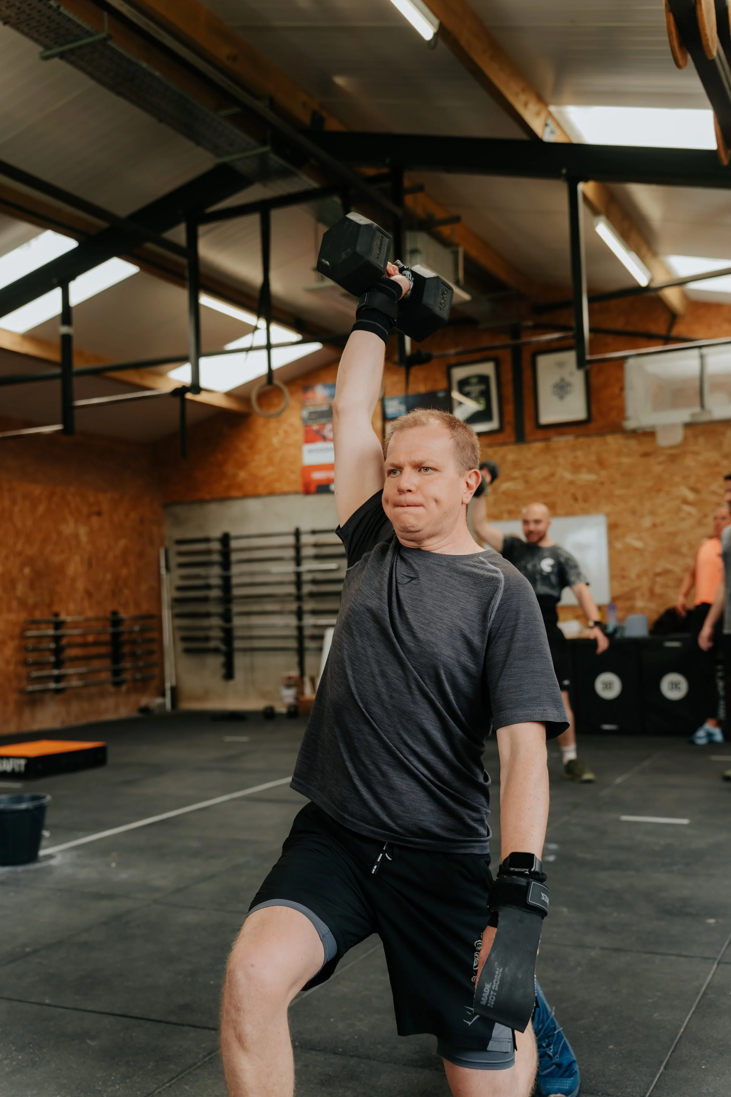 A man lifting a dumbbell in a gym with other people in the background.