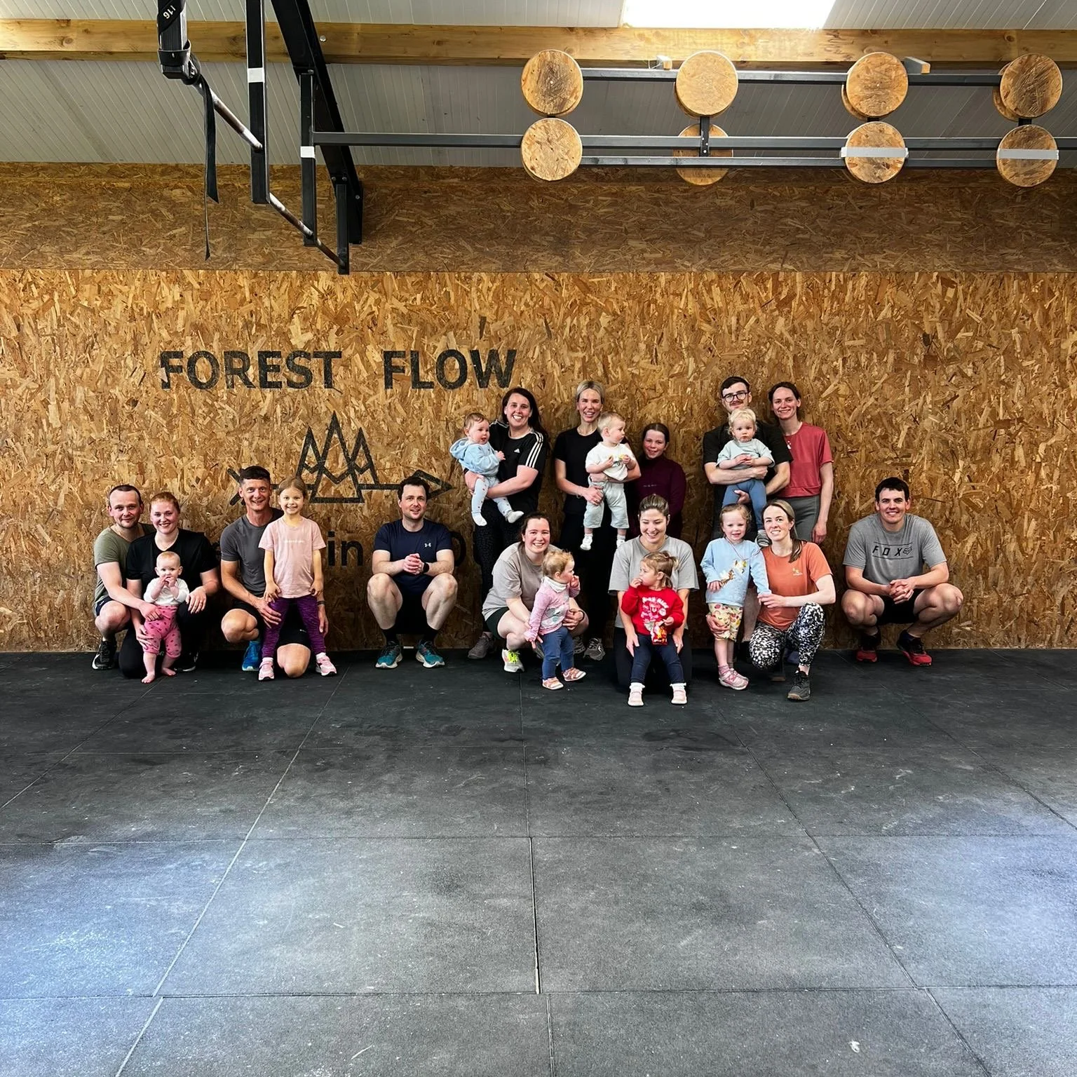 A group of adults and children smiling and posing for a photo in an indoor gym or fitness facility with a wooden wall behind them and the words 'FOREST FLOW' written on the wall.