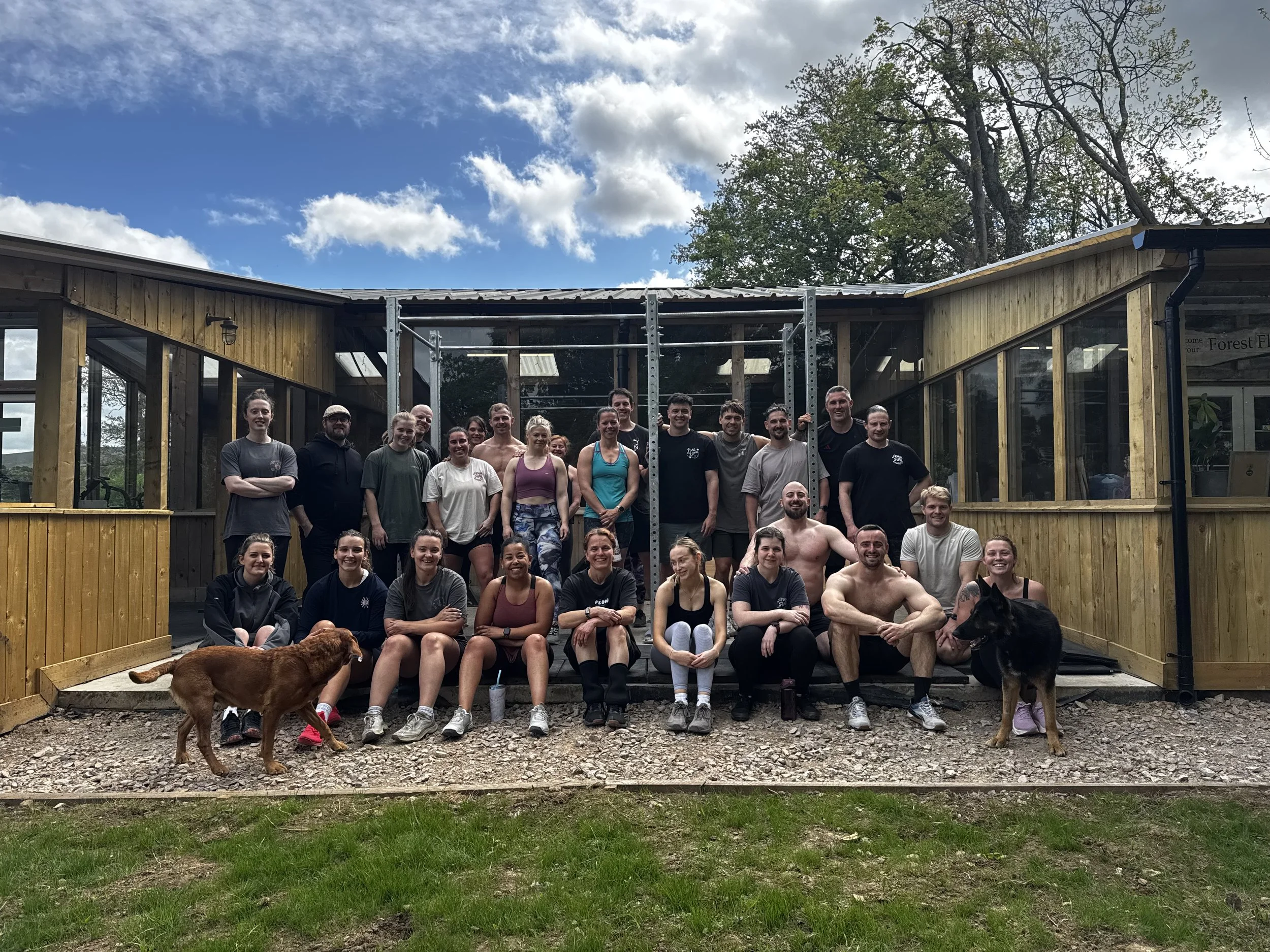 A large group of people, including men and women, are gathered outdoors in front of a wooden building with glass windows, under a partly cloudy sky. Some are sitting, some are standing, and a couple of dogs are present in the scene.