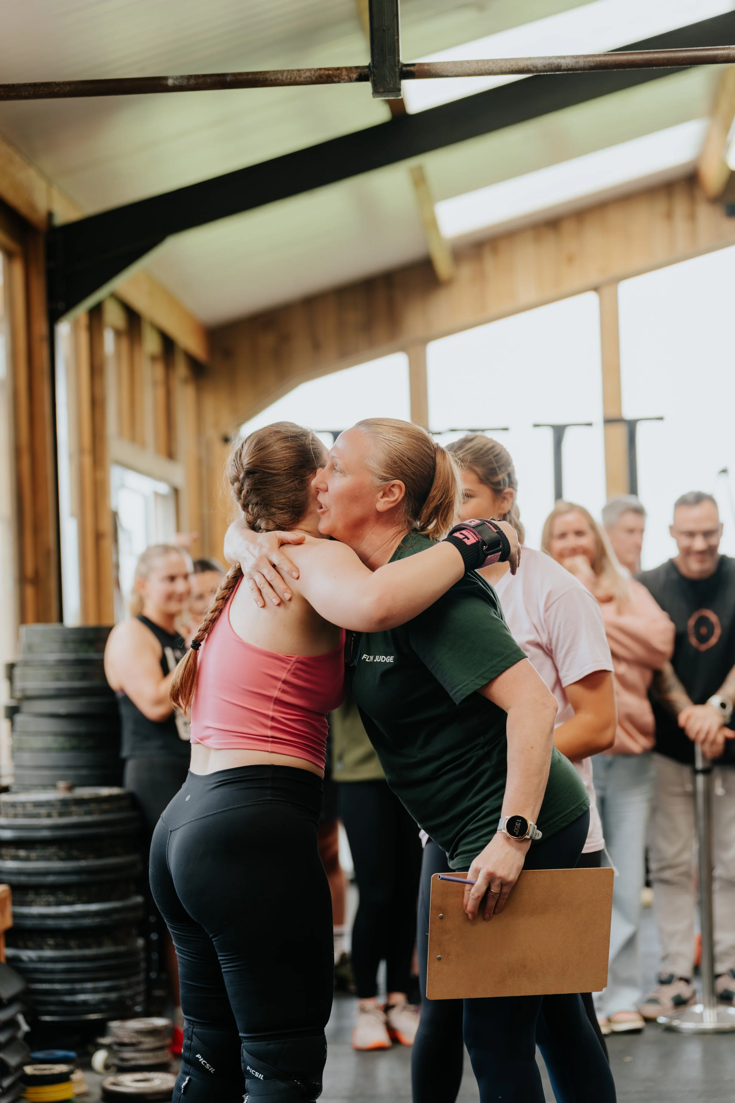 Two women embrace in a gym setting, surrounded by others, with workout equipment in the background.
