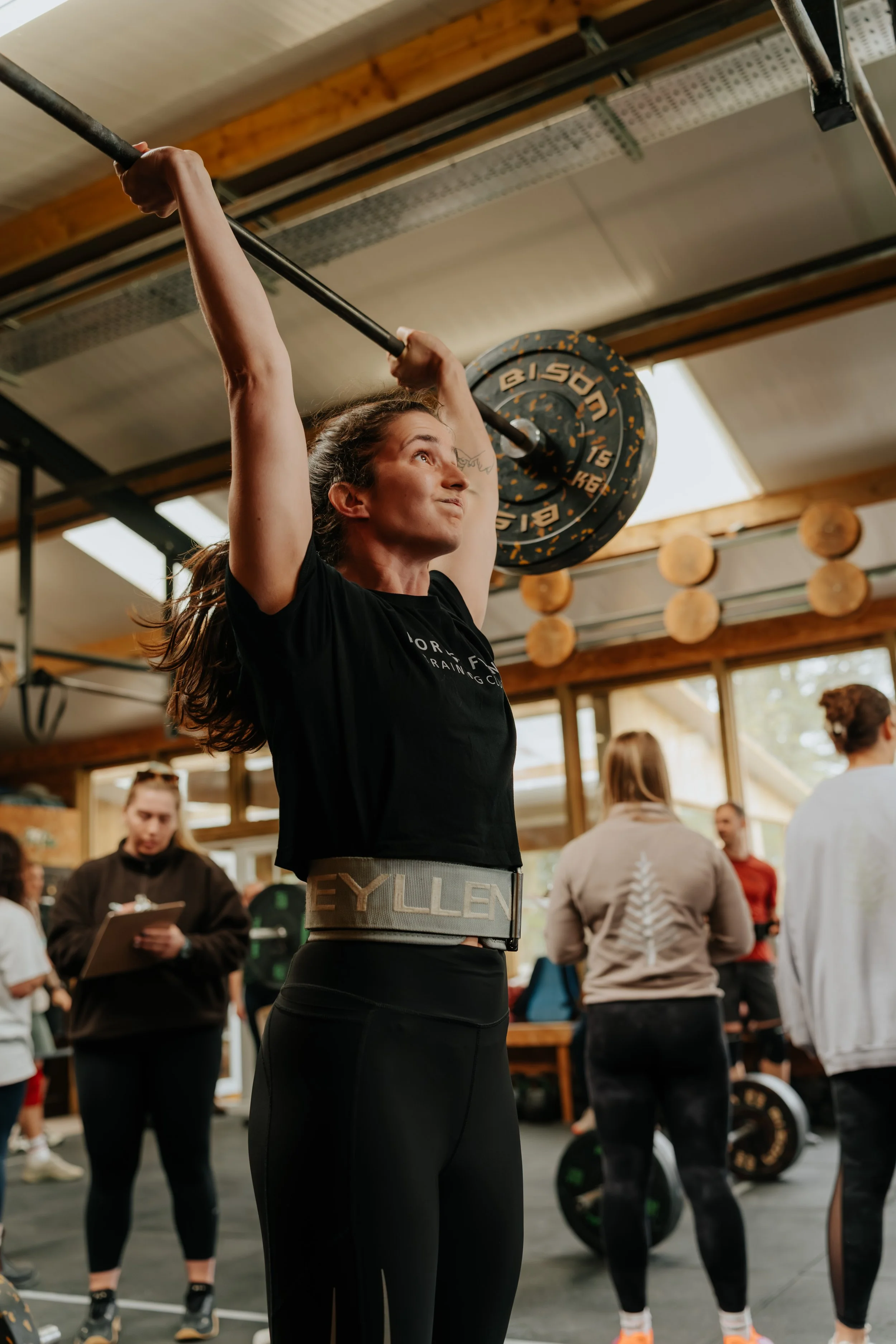 A woman lifts a barbell at a gym, surrounded by several people, with wooden beams and large windows in the background.