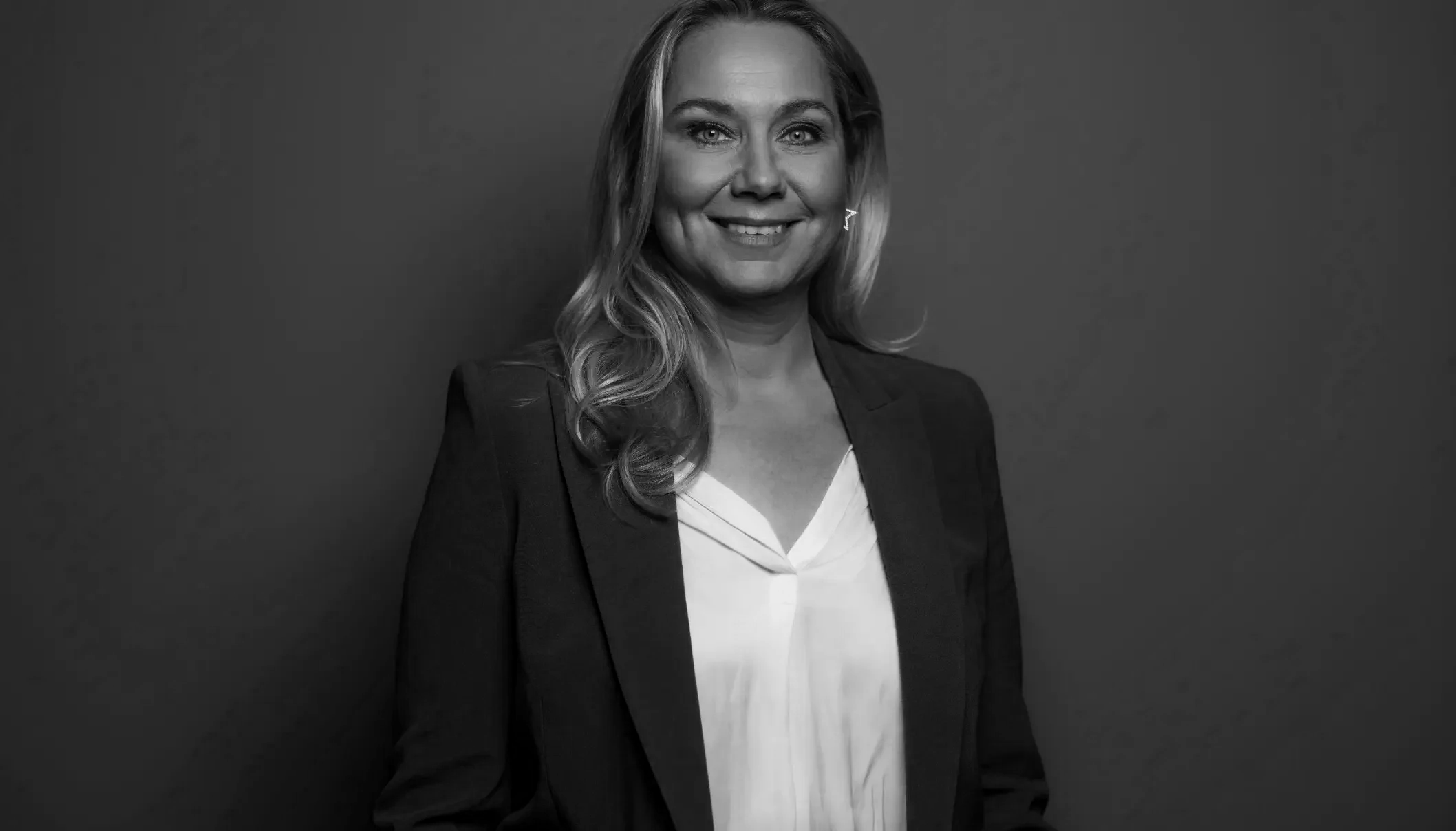 Black and white portrait of a smiling woman with shoulder-length hair, wearing a blazer and a blouse, against a plain wall background.