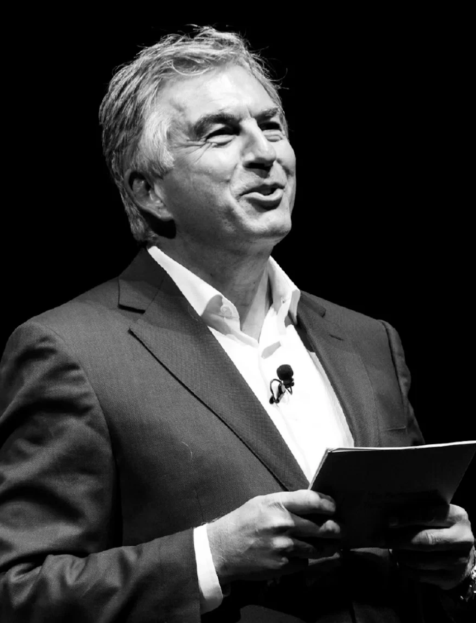 A black-and-white photo of a man in a suit and white shirt, smiling and holding a tablet while speaking or presenting.