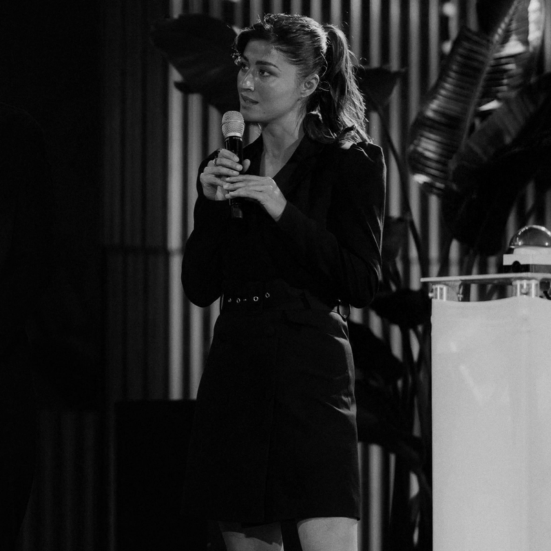 A woman with dark hair tied back, wearing a dark blazer and skirt, holding a microphone and speaking on stage with vertical curtains and large potted plant in the background.