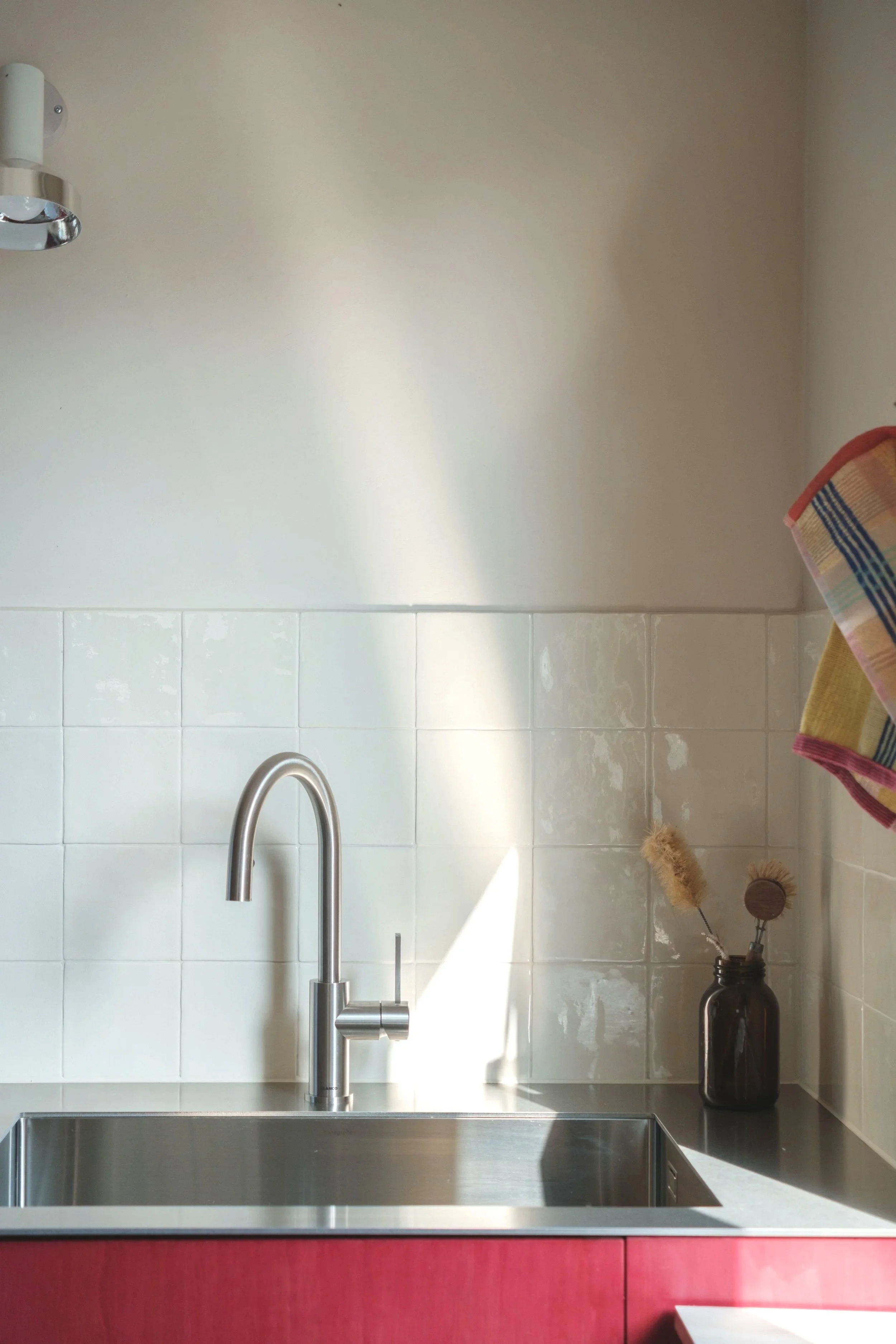Moderner Küchenspüle mit Edelstahlbecken, Wasserhahn, weiße Fliesen an der Wand im Hintergrund, dunkler Küchenunterschrank, Dekokorb mit Trockenblumen, Handtuch hängt an der Wand.