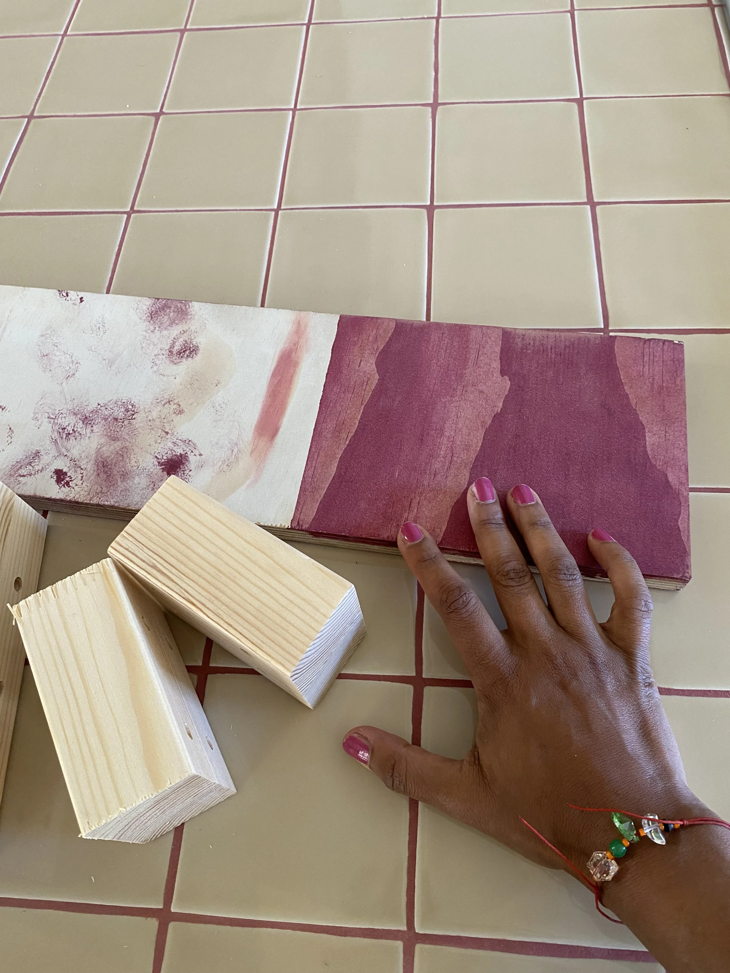 Eine Hand, die auf einer Oberfläche mit roten Kacheln liegt, hält ein farbiges Buch oder eine Zeitschrift mit einer blumigen und einem rot-gestreiften Muster. Daneben liegen Holzteile.