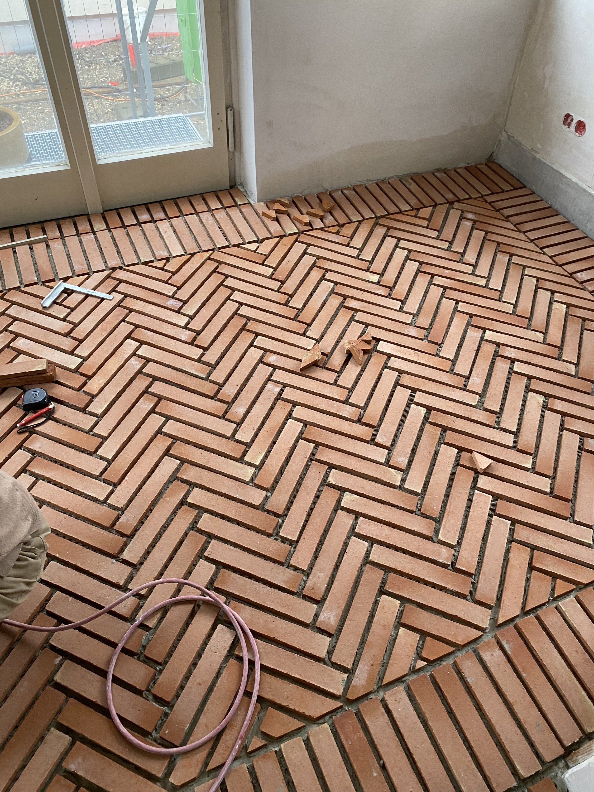 Der Boden ist mit roten Ziegelsteinen in einem diagonalen Muster verlegt, und es sind noch einige lose Ziegelsteine auf dem Boden. Es gibt auch ein Messgerät und ein Kabel im unteren Bereich des Bildes. Das Bild zeigt einen Innenraum während der Baup