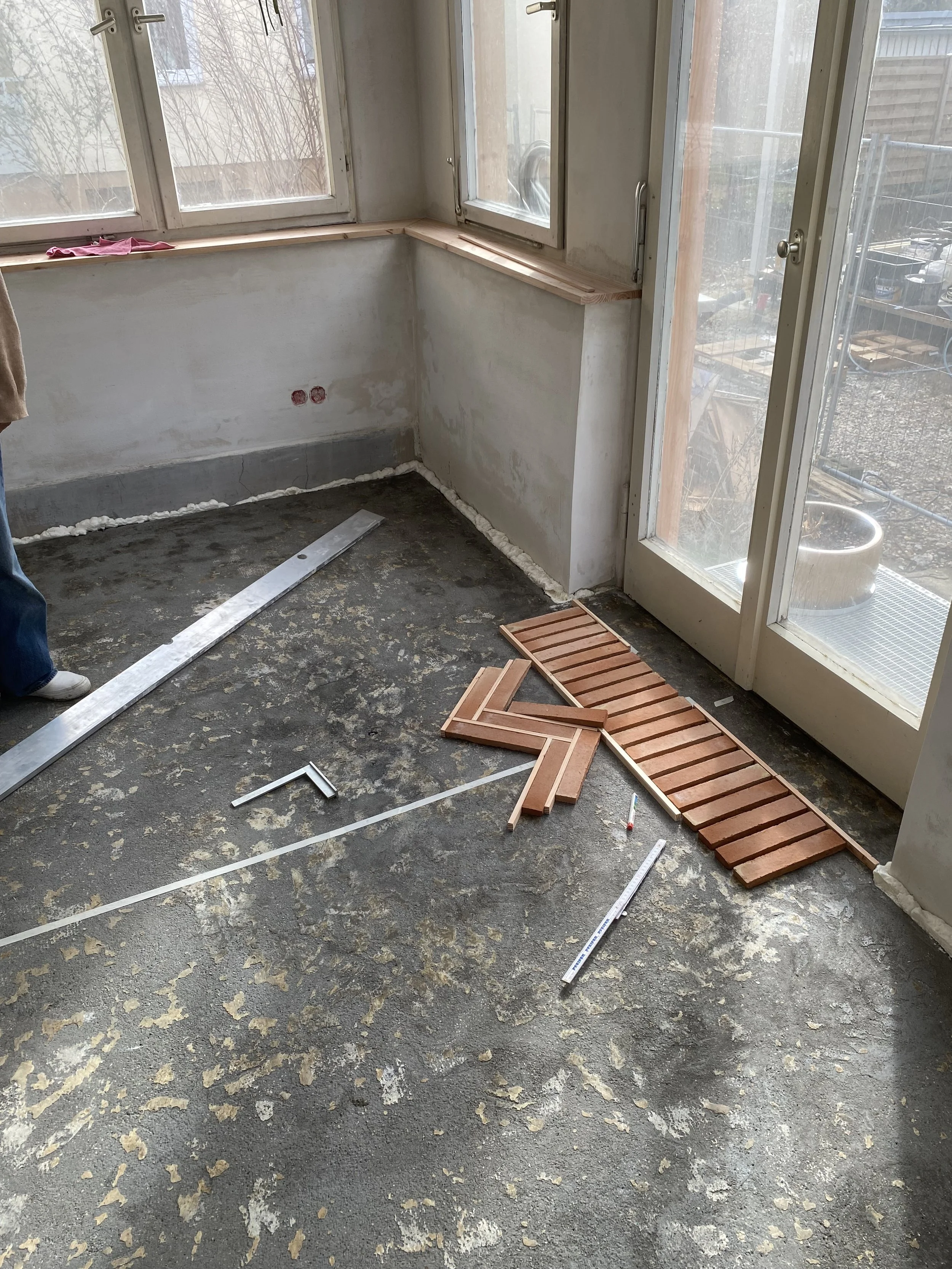 Ein Raum im Bau mit einem ungefassten Boden, einer weißen Wand mit roten Kabelanschlüssen, mehreren Messwerkzeugen auf dem Boden, eine Tür mit Glas und eine Fensterfront. Es wird wahrscheinlich an der Renovierung oder dem Bau einer Terrasse oder eine
