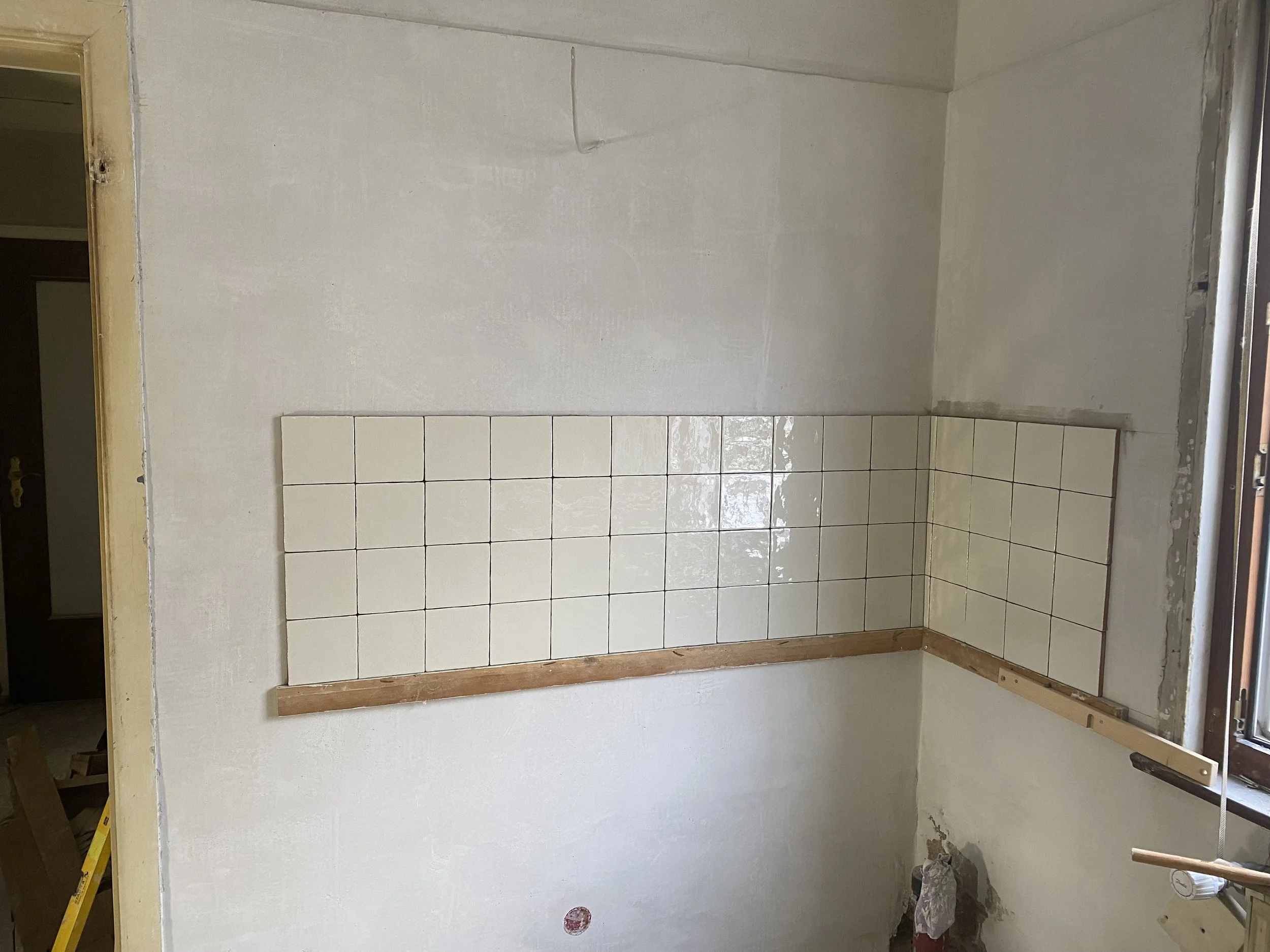 Un fertiggestellter Raum mit weißen Wandfliesen an einer Wand, die noch verputzt wird, und eine kleine Fensteröffnung. Der Boden ist noch nicht fertiggestellt, und es sind Baumaterialien und Werkzeuge sichtbar.