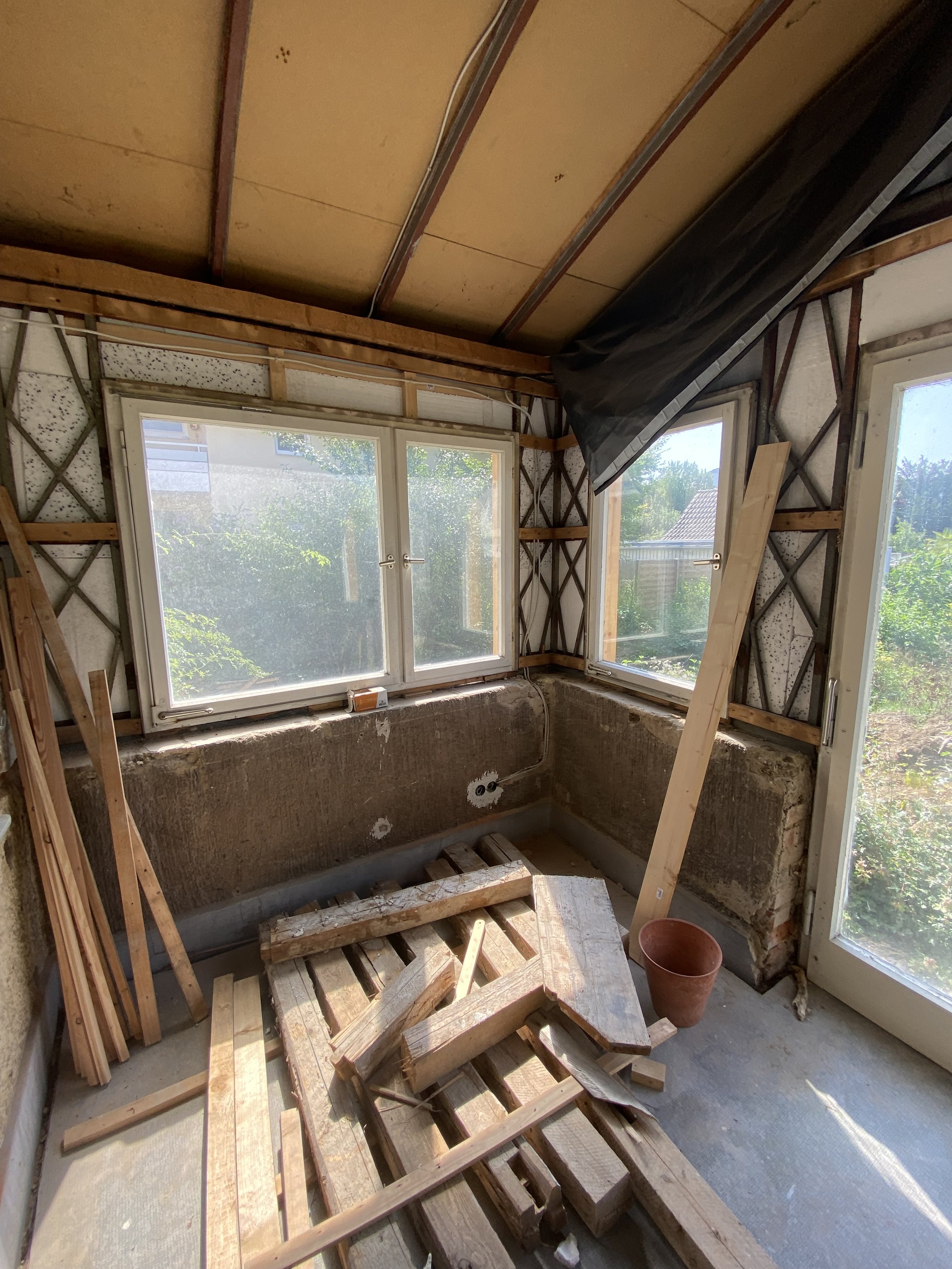 Ein Raum im Aufbau mit Fenstern, unvollständige Wände, Baumaterialien und Holzreste auf dem Boden, wahrscheinlich eine Renovierung oder Bauarbeiten im Gange.