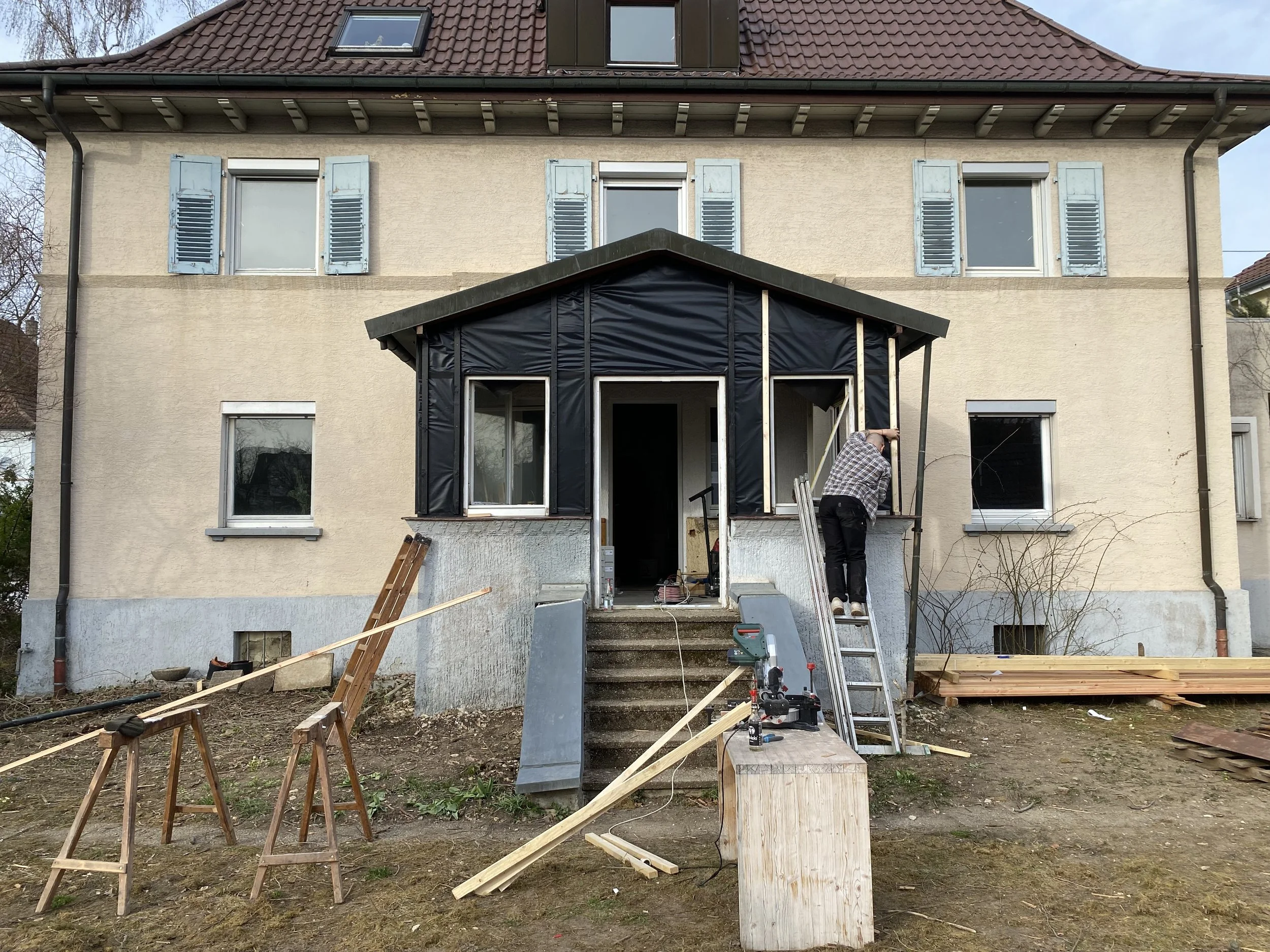 Ein Mehrfamilienhaus wird renoviert oder gebaut, mit einem Baugerüst, einer Leiter und Baumaterialien im Gartenbereich vor dem Haus.