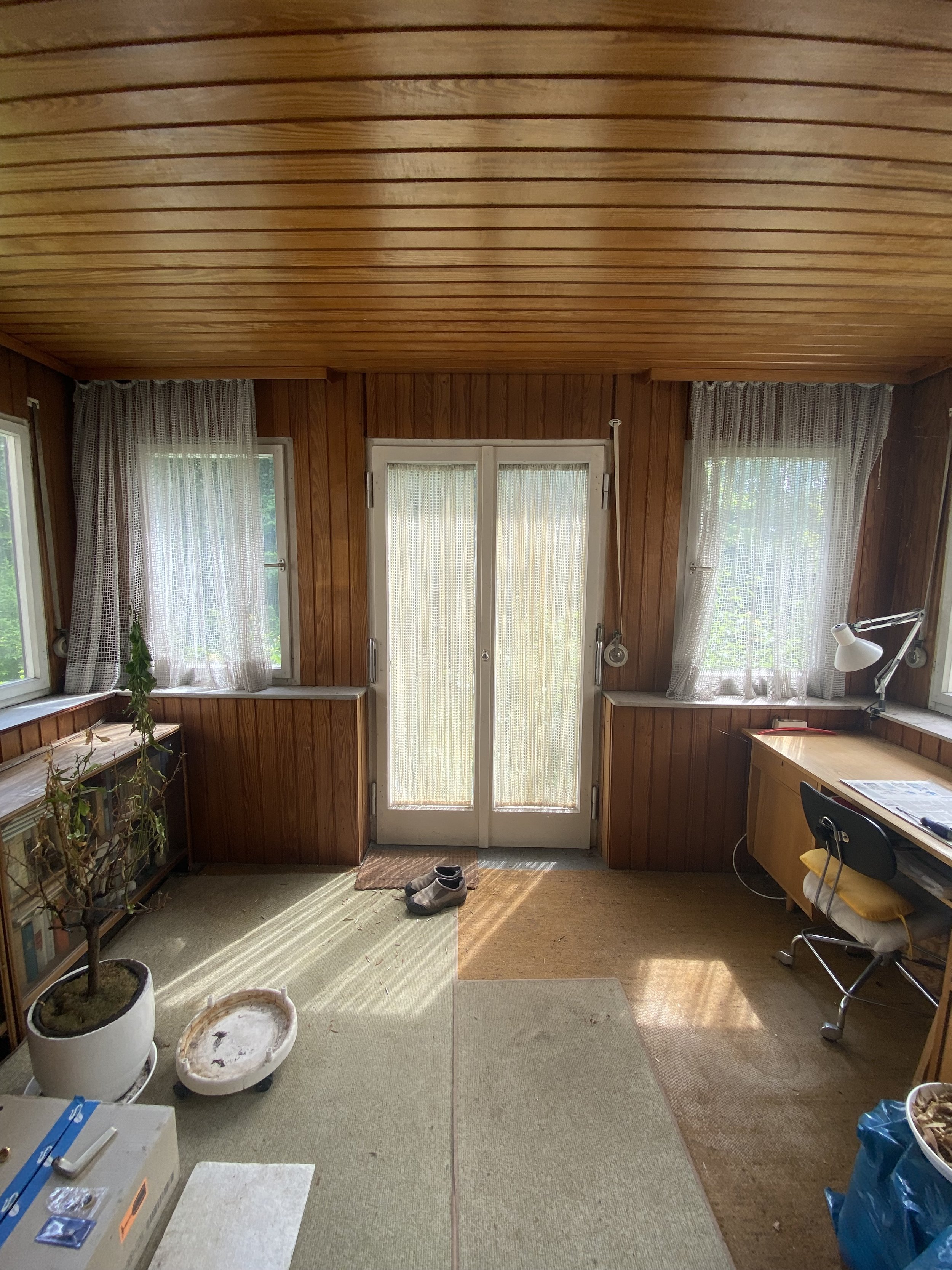Ein Raum mit Holzpaneelen an Wänden und Decke, mehrere Fenster mit Vorhängen, Eingangstür mit Glas, ein Schreibtisch mit Lampe, Schuhe vor der Tür, Pflanzen und verschiedene Gegenstände im Raum.