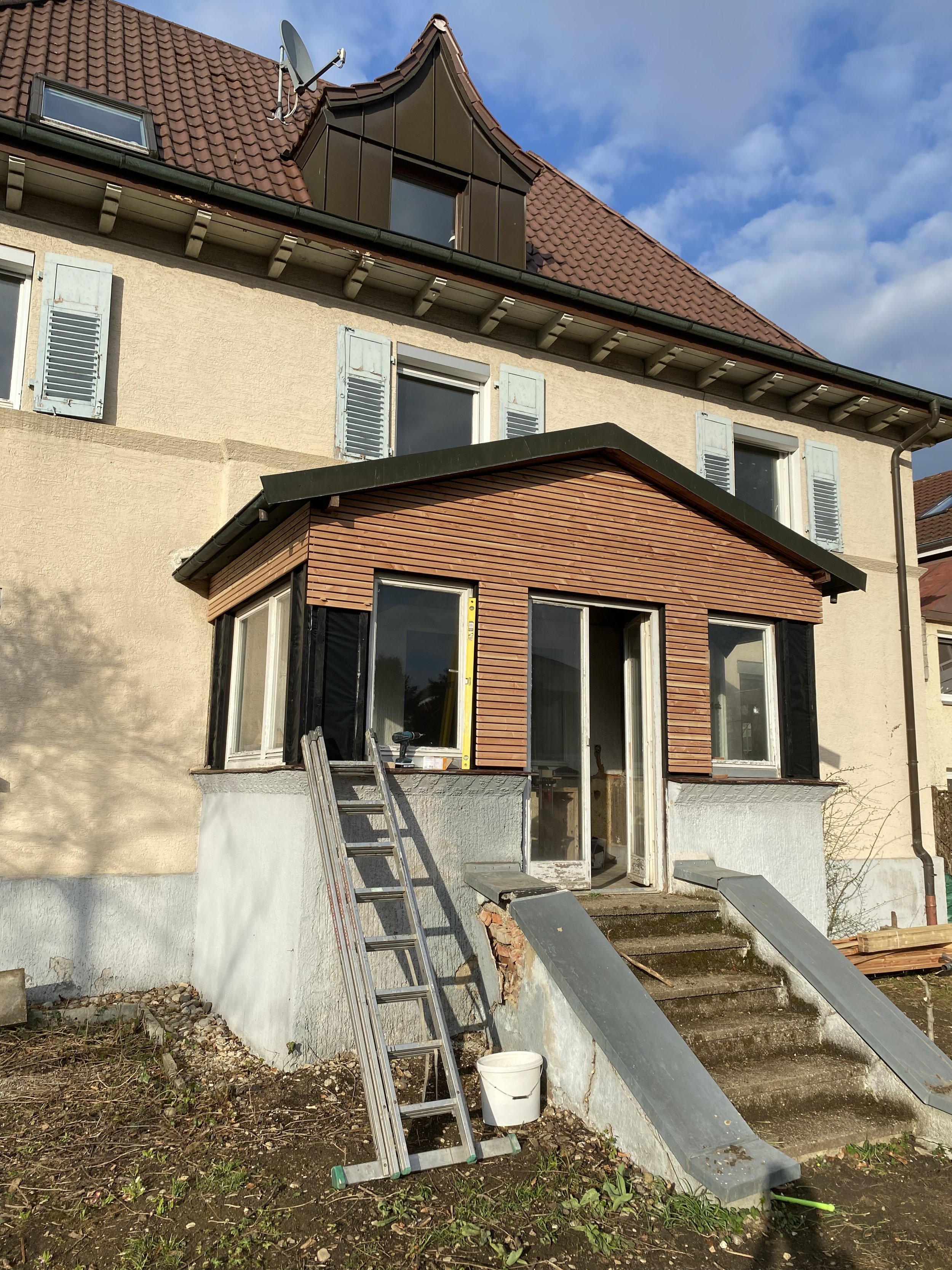 Ein Haus wird derzeit renoviert oder gebaut. Es gibt eine Leiter, Baumaterialien und eine verglaste Terrasse im Garten.