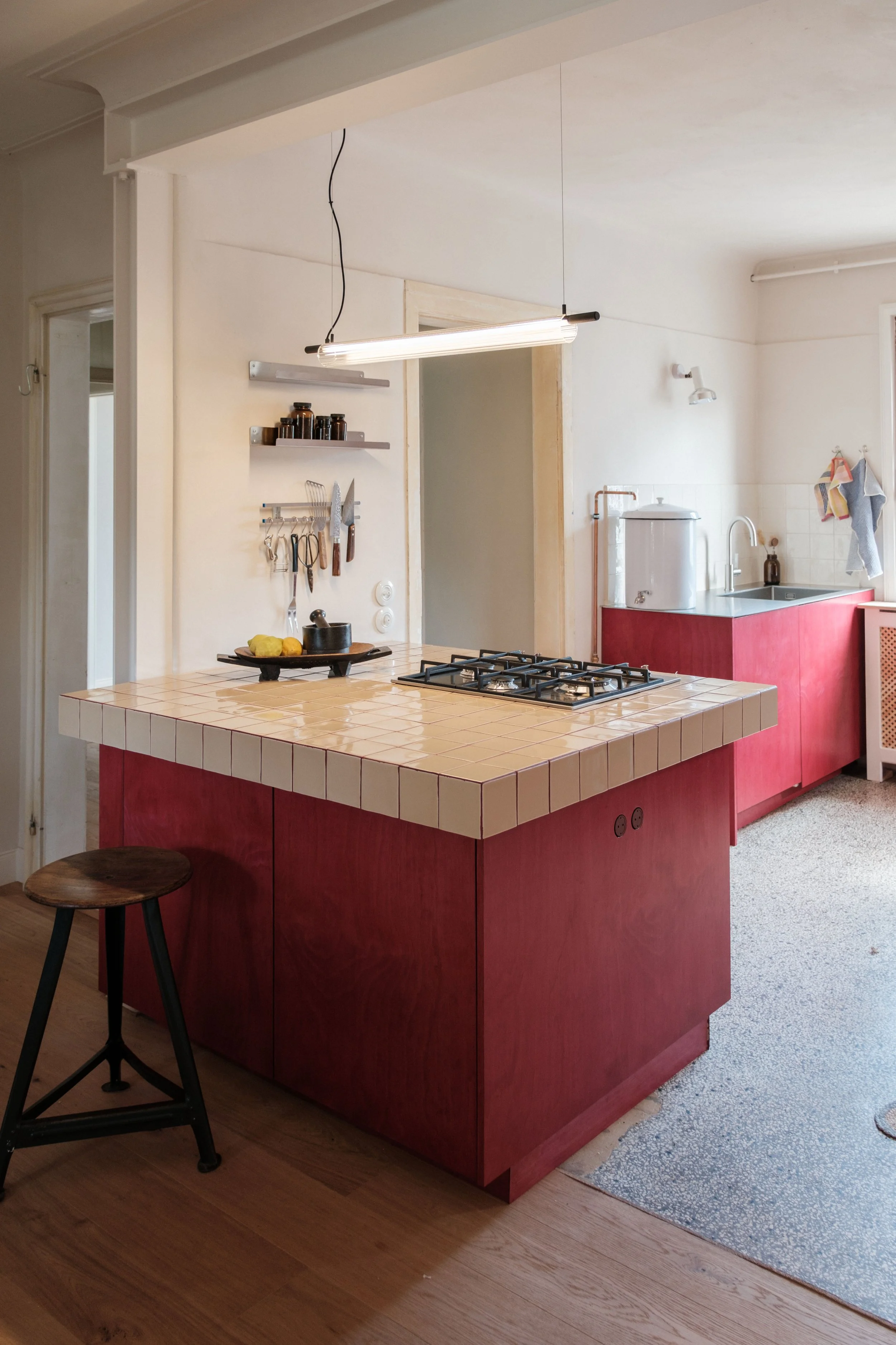 Eine Küchentheke mit roten Türen, einer lila Mikrowelle, einem Wasserkocher, einem Wasserhahn, und Handtüchern am Fenster. Eine Holzhocker steht neben der Theke.