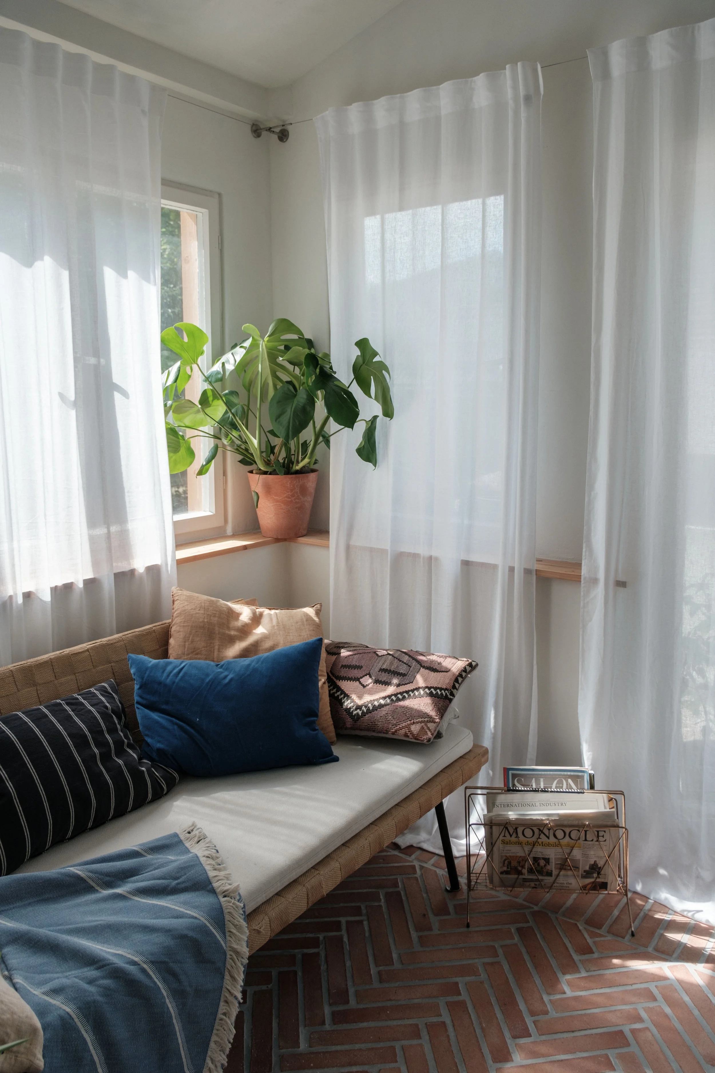 Ein Wohnraum mit weißen Vorhängen am Fenster, einer Pflanze in einem Terrakottatopf auf dem Fensterbrett, und einer Couch mit verschiedenen Kissen sowie einem Korb mit Zeitschriften.