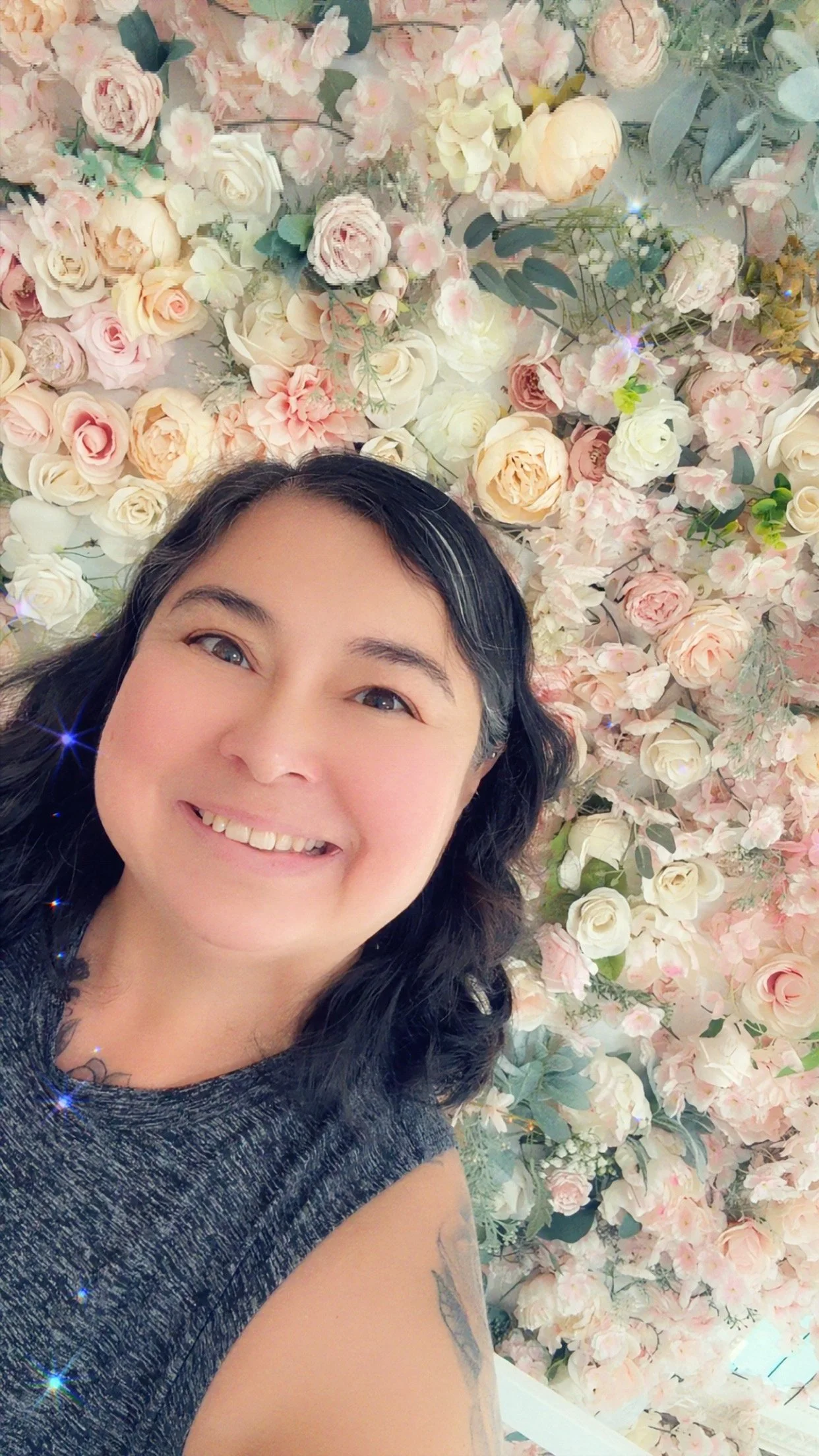 A woman smiling in front of a wall of pink and white flowers.
