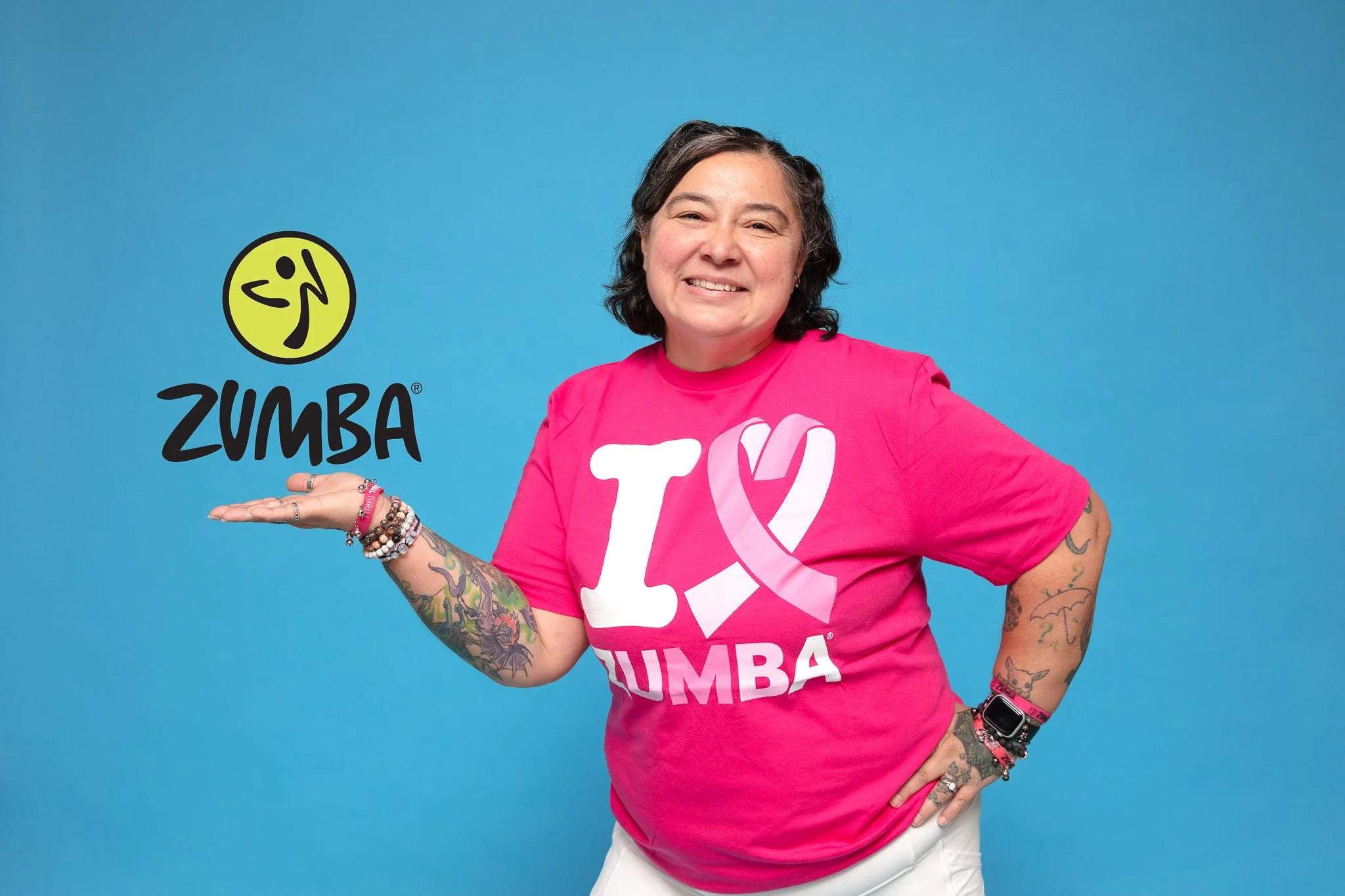 A woman with tattoos on her arms, wearing a bright pink T-shirt with a white and pink logo, smiling and posing with her right hand extended. She stands against a solid blue background.