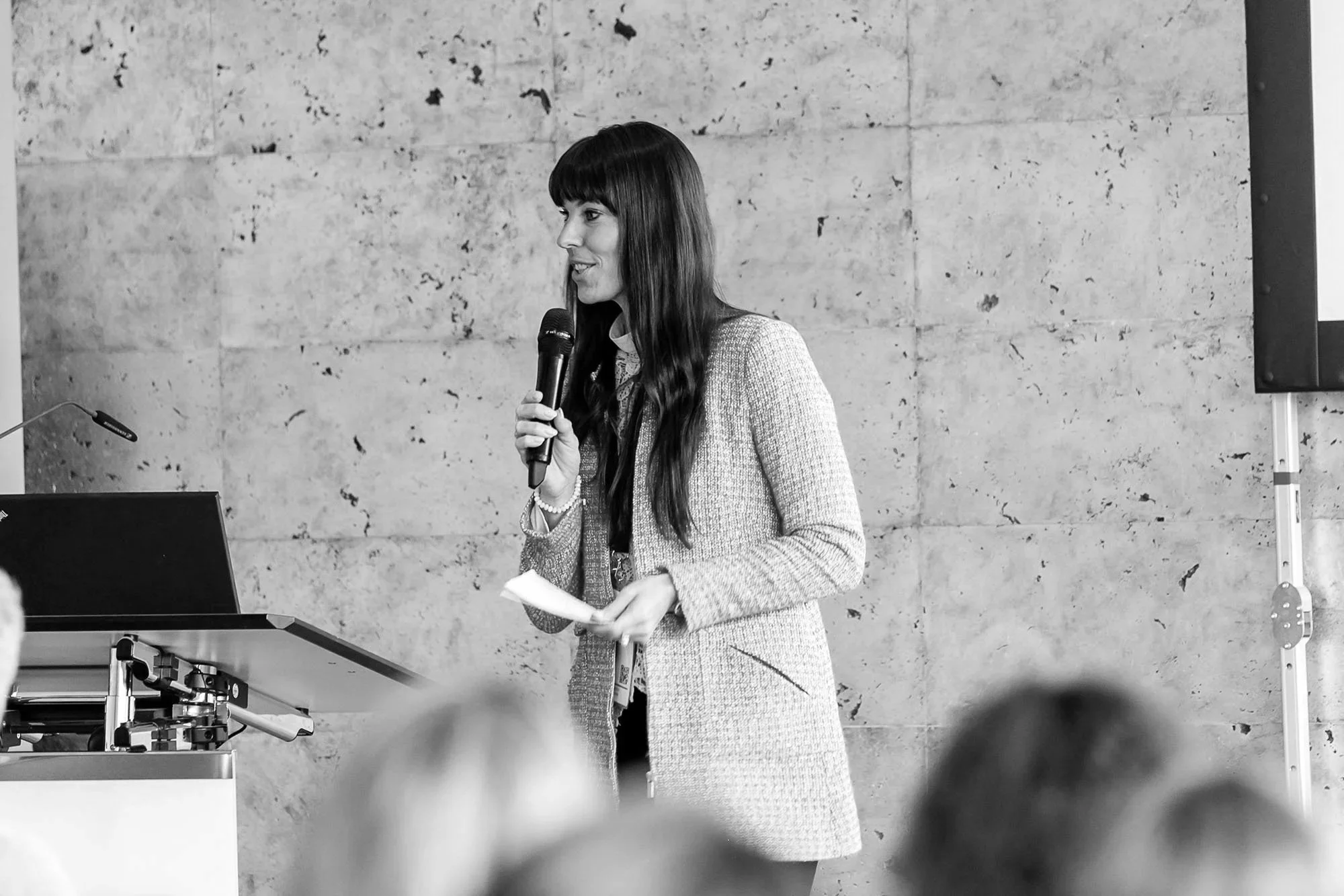 Sarah Johannsen Roth spricht in einem Raum mit Betonwand, hält ein Mikrofon und liest Notizen, während ein Podium und technische Geräte im Vordergrund sichtbar sind.