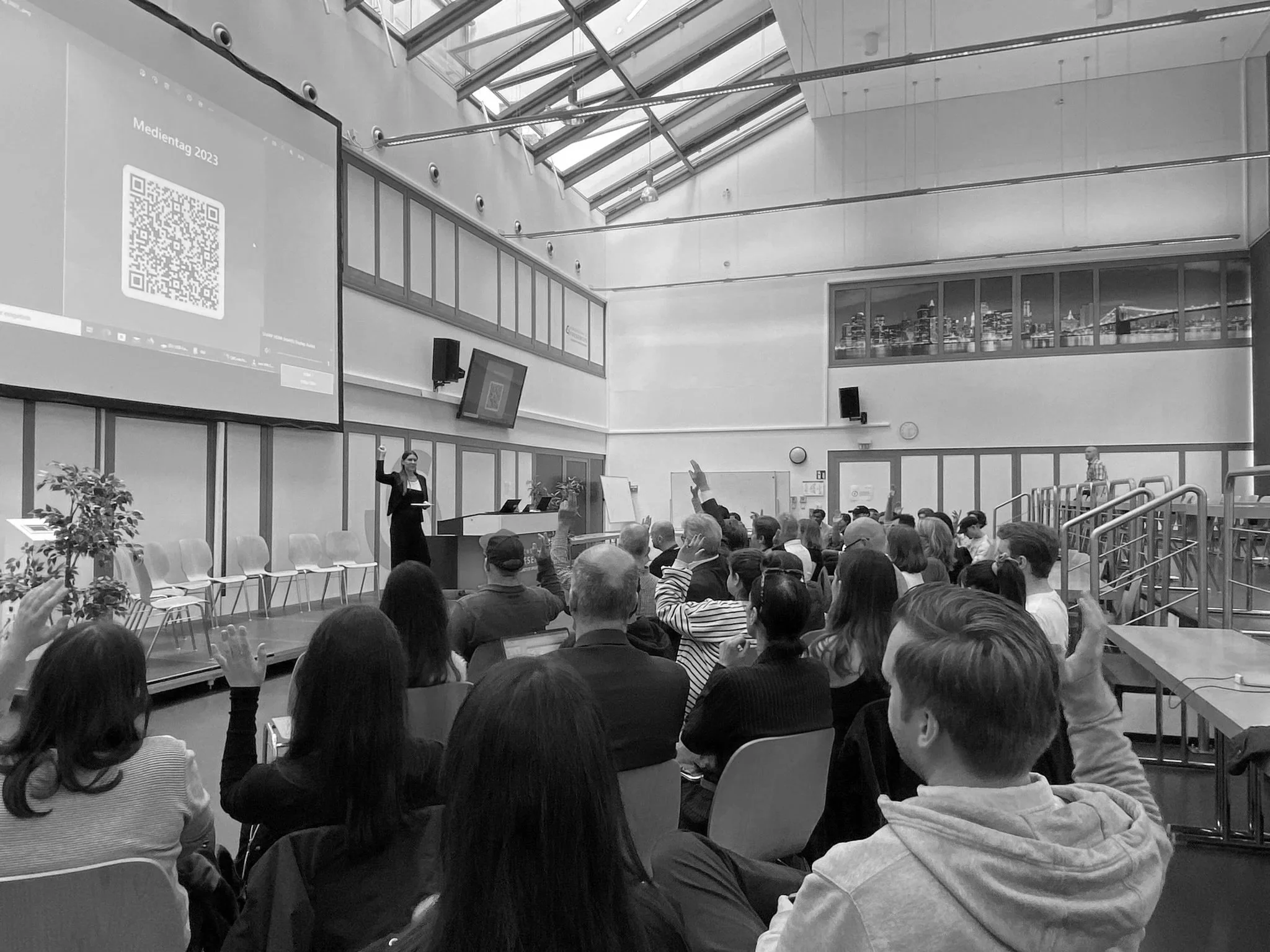 Eine große Gruppe von Menschen sitzt in einem modernen Konferenzraum mit hohen Decken und großen Fenstern. Es findet eine Präsentation statt, wobei eine Frau an der Seite des Raumes steht und spricht. Einige Zuhörer heben die Hände, wahrscheinlich um Fragen zu stellen oder abstimmen. Im Hintergrund sind große Monitore und eine Projektionsfläche sichtbar, auf der farbige QR-Codes und Text gezeigt werden.