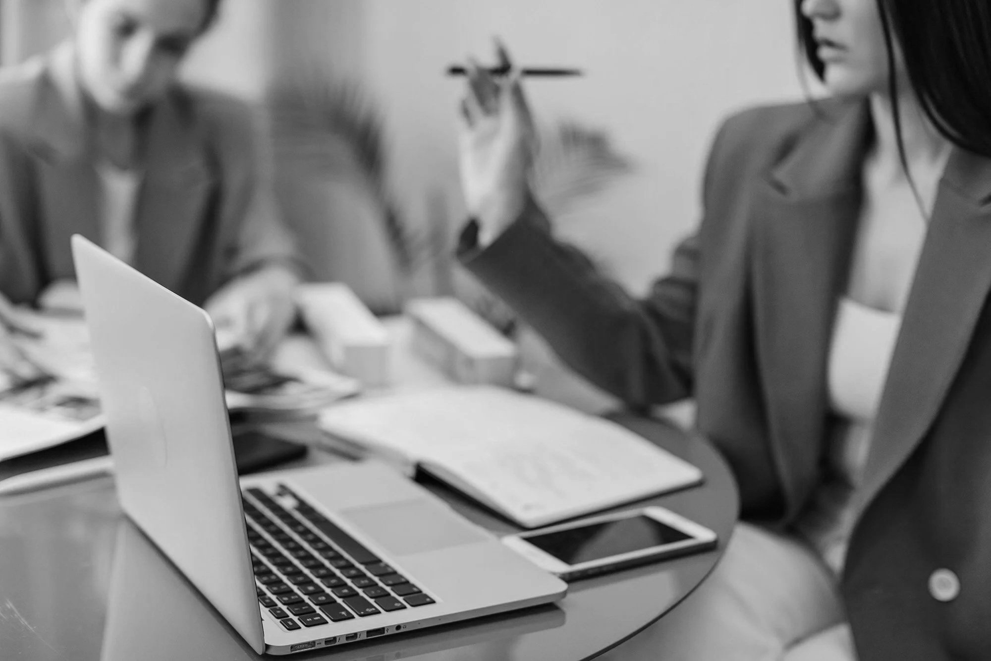 Zwei Frauen arbeiten in einem Büro an einem Tisch, neben einem Laptop. Eine Frau trägt eine Jacke und hat dunkle Haare, die andere ist leicht sichtbar und trägt ein weißes Hemd.
