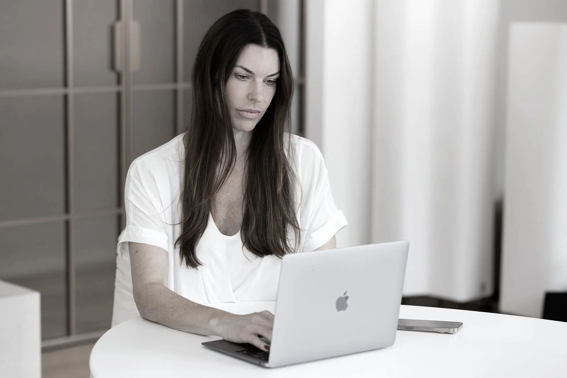 Sarah Johannsen Roth arbeitet an einem weißen Tisch mit einem weißen Laptop in einem modernen, hellen Büro. Sie trägt ein weißes T-Shirt und scheint konzentriert zu sein.