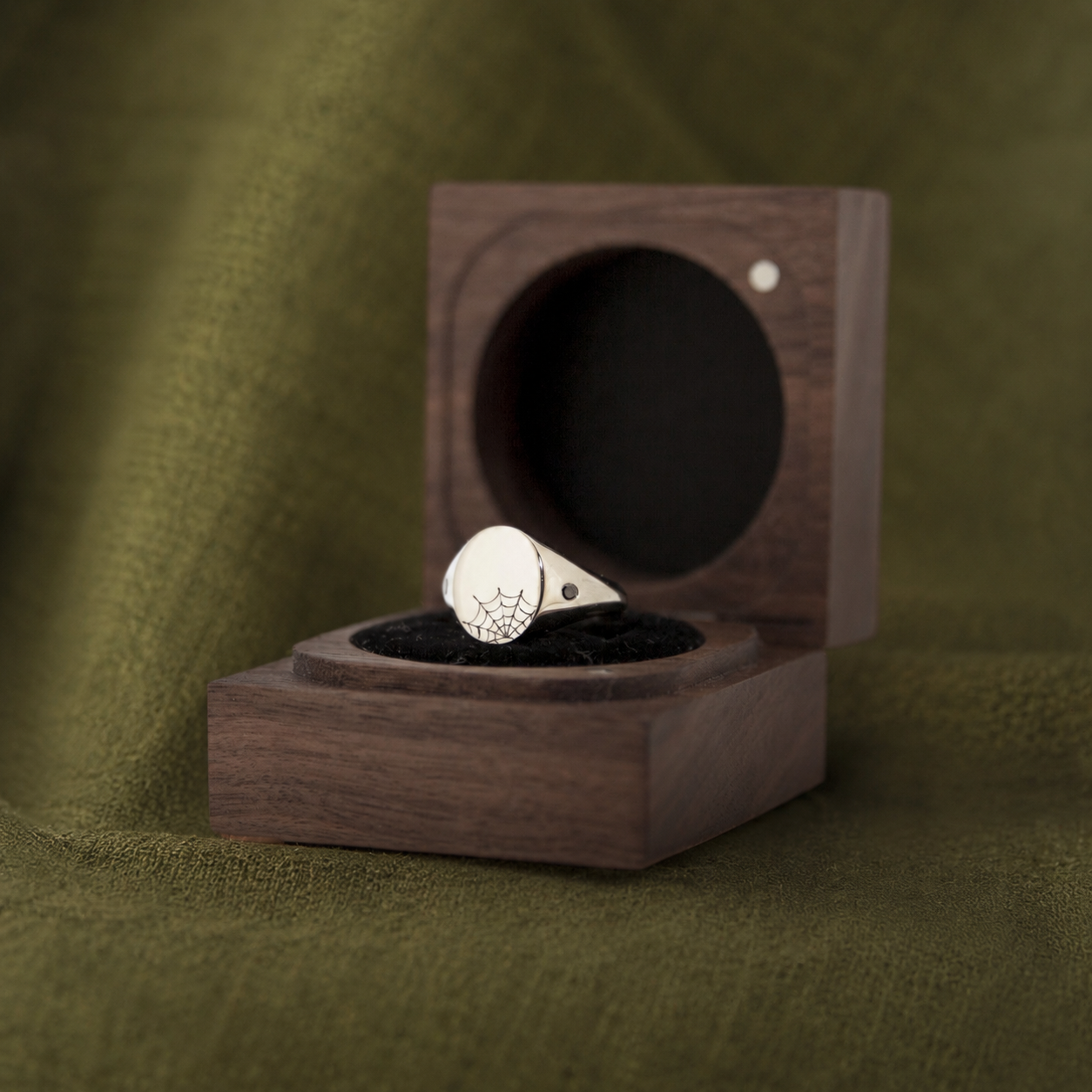 A silver ring with a spider web design on top, placed inside an open wooden ring box with a black velvet lining, on a green textured fabric background.