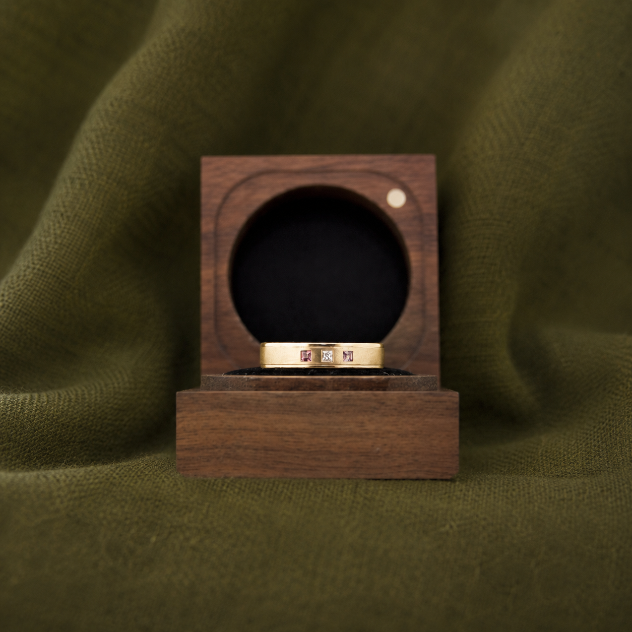 A gold ring with gemstones sits on a wooden display stand, with a green fabric background and a wooden box with a circular opening behind it.