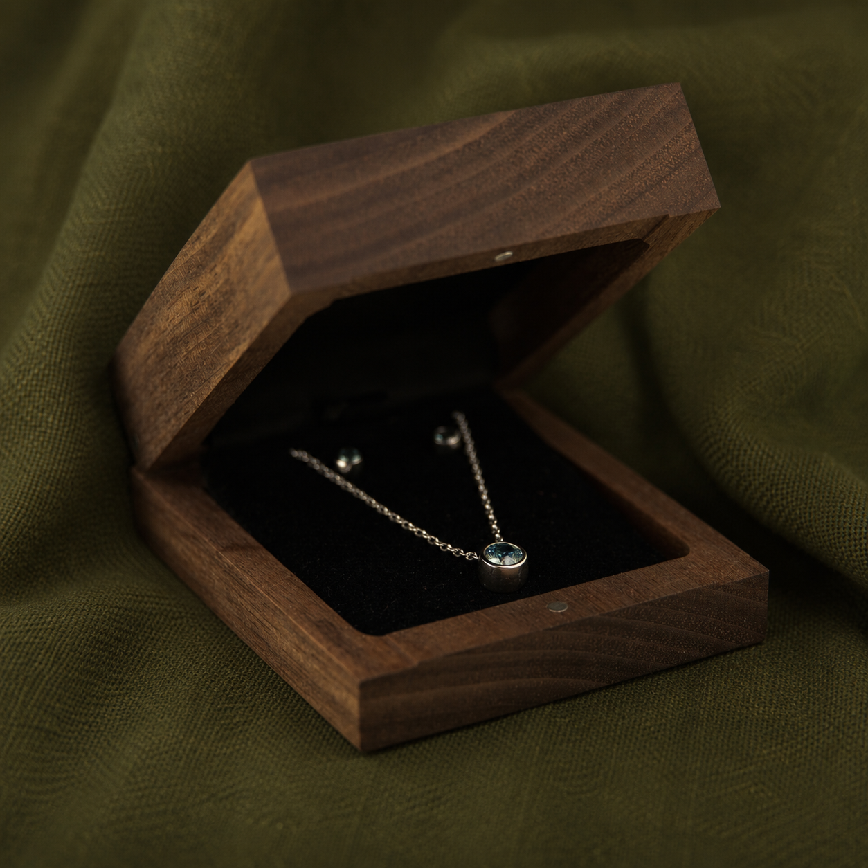 A necklace with a blue gemstone pendant and matching earrings inside a wooden jewelry box, set on green fabric.