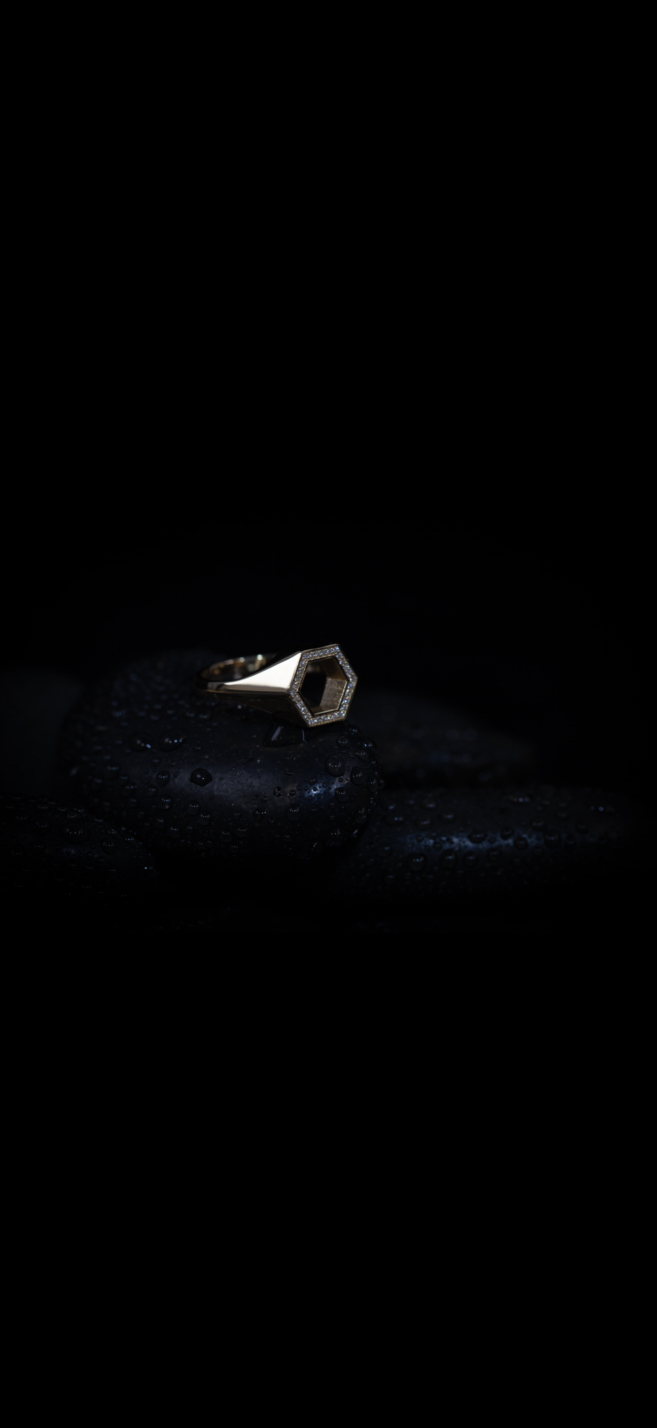 A silver ring with a hexagon-shaped top, surrounded by small diamonds, rests on black stones with water droplets.