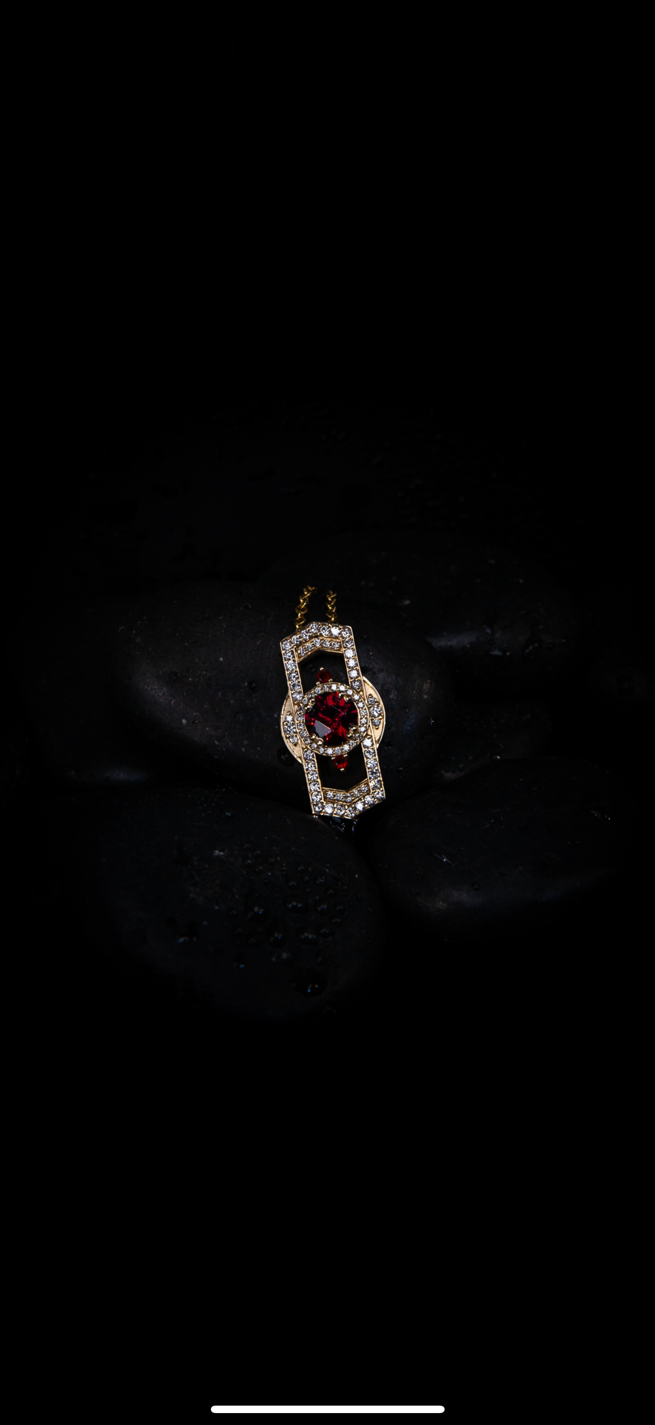 A gold necklace with a pendant featuring red and clear gemstones, resting on black stones with water droplets.