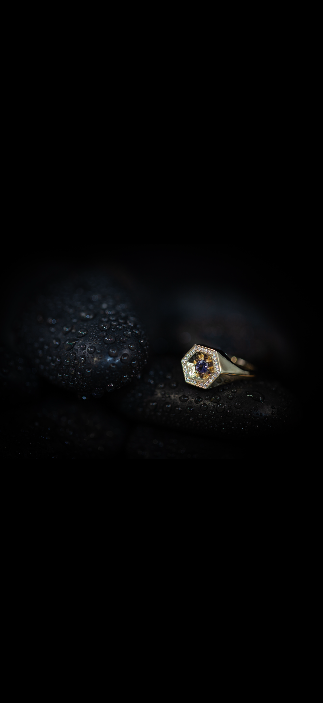 A gold ring with a central blue gemstone surrounded by smaller diamonds on smooth, wet black stones.