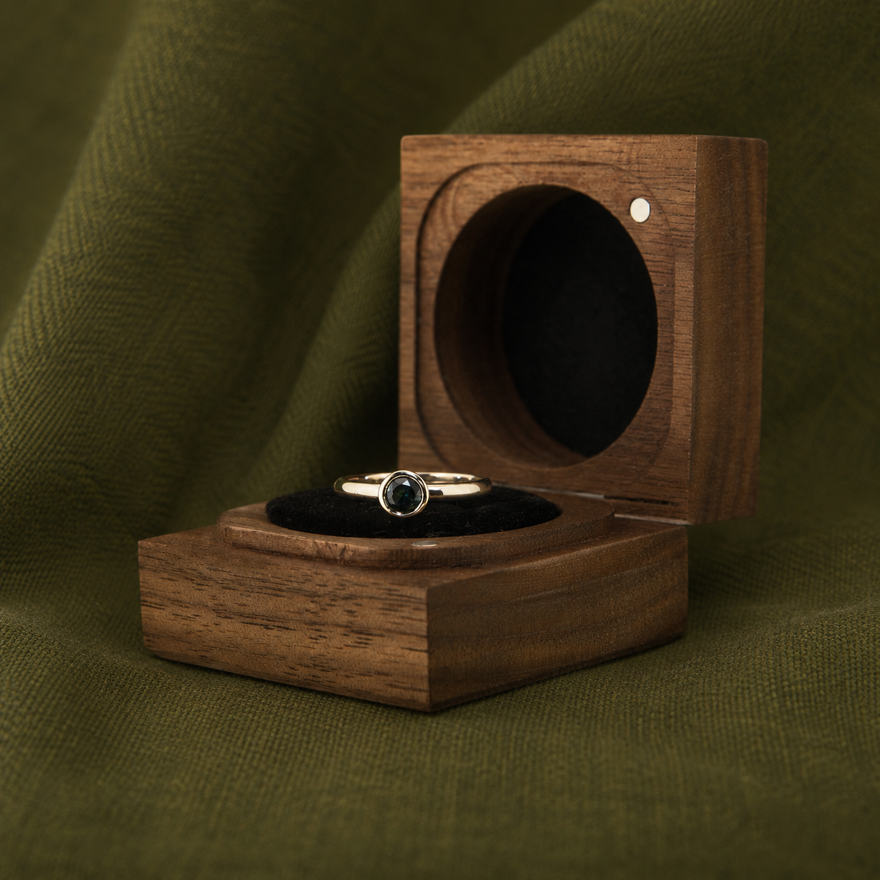 Silver ring with a black gemstone placed inside an open wooden ring box, set against a green fabric backdrop.