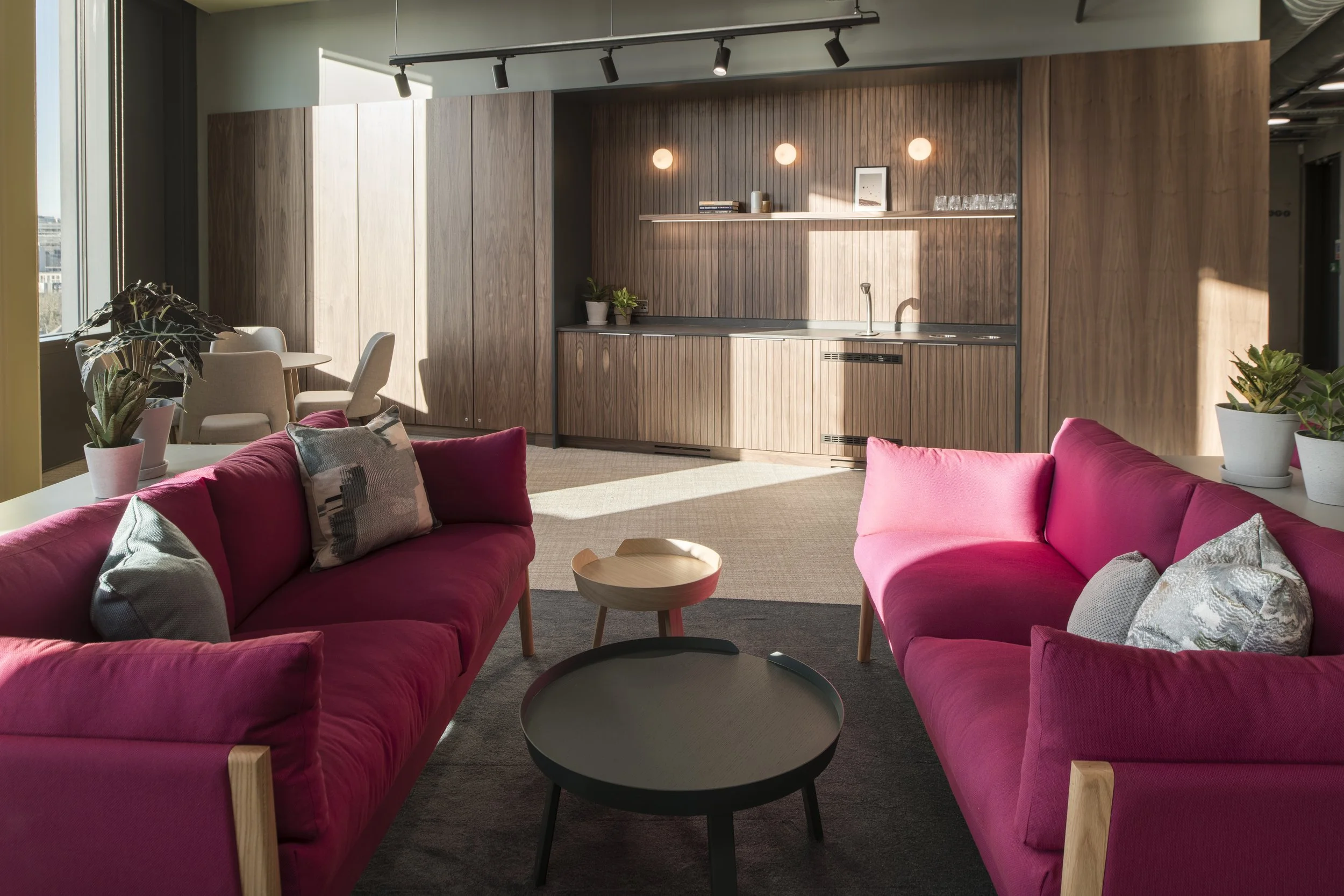 Modern office lounge with pink sofas (Boss Bodie Sofa), Muuto around coffee tables, and plants. Bespoke joinery design, walnut panelling and bespoke office kitchen design. Workplace interior design Bristol. Clarke Willmott Solicitors Bristol Assembly