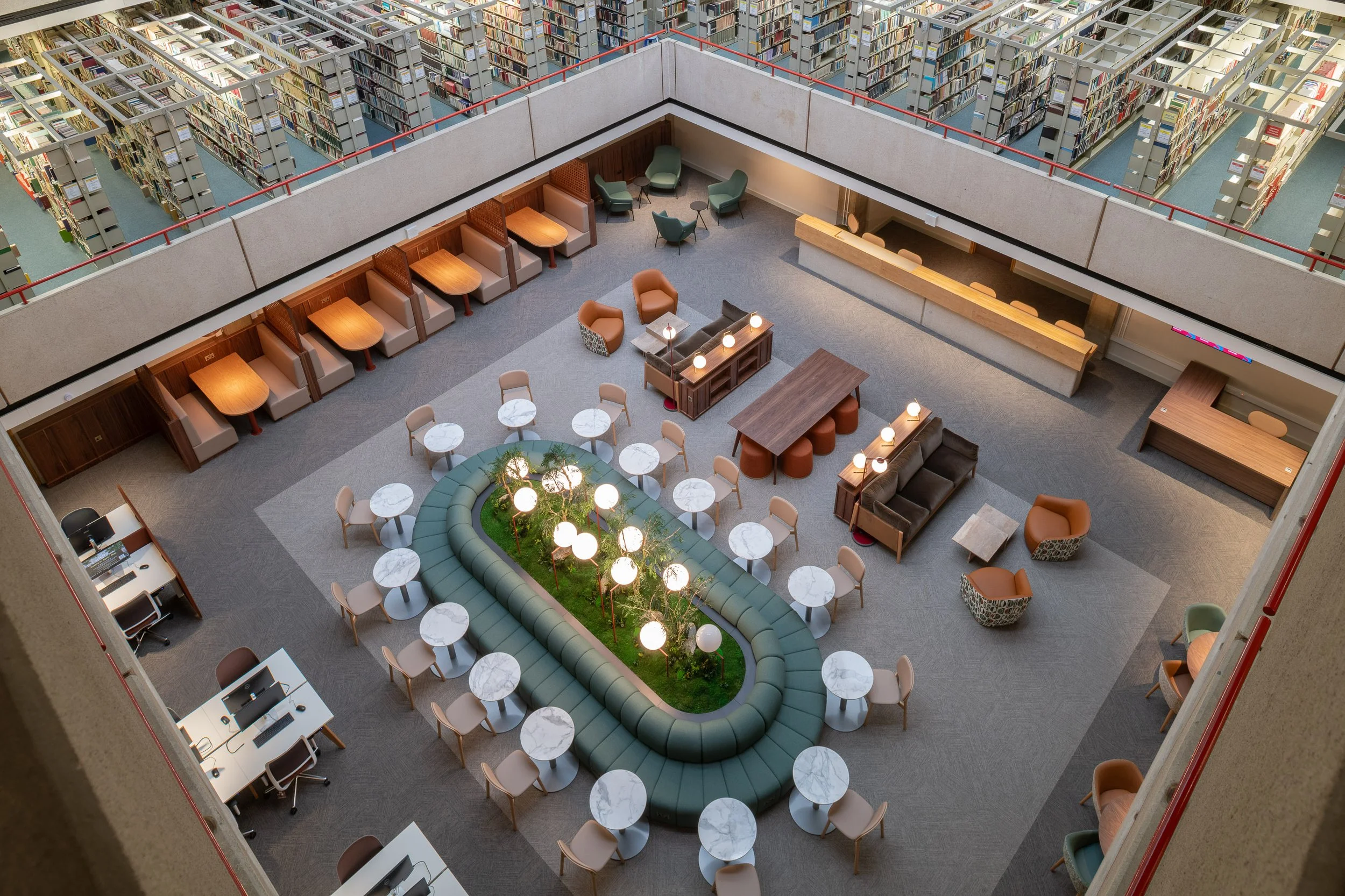 SOAS University of London Library Interior Design. A view of a modern library interior looking down from the upper floor of a brutalist atrium space. Mood lighting, lounge seating, and a circular green banquette with indoor planting.
