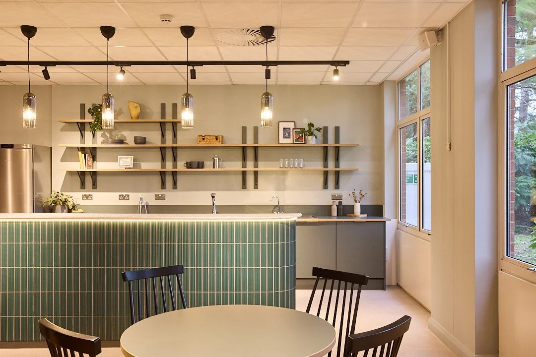 Modern café interior design with green tiled counter, open wooden shelves, pendant lights, round dining tables, black ercol chairs and large windows. Midcentury style. Workplace kitchen fit out Bristol. Office breakout area interior design Bristol.