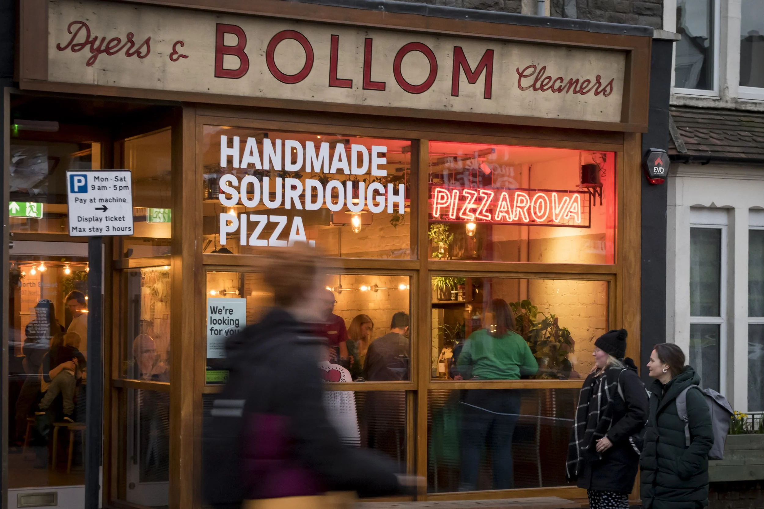 Exterior of Pizzarova Bristol North Street shop front design. A crowed pizza restaurant with timber sash windows, and a glowing neon sign. A ghost sign above says 'Bollom'. Restaurant interior design Bristol. Bespoke shopfront design.