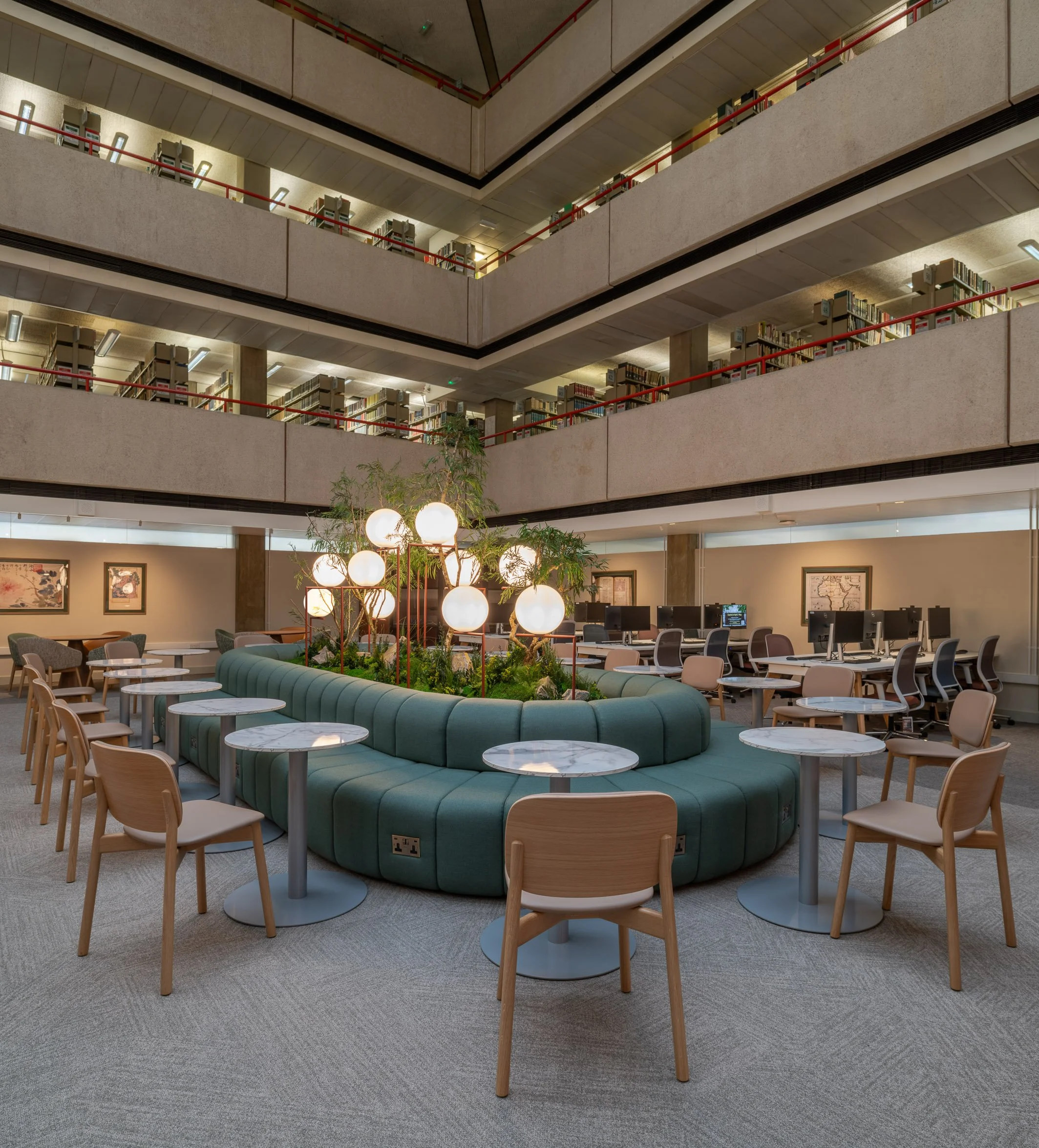 Modern multi-level library workplace interior design with bookshelves, reading tables, computers, and a circular green seating area around a plant installation. Bespoke central banquette seating, round dining tables, and Boss kitt dining chair oak.