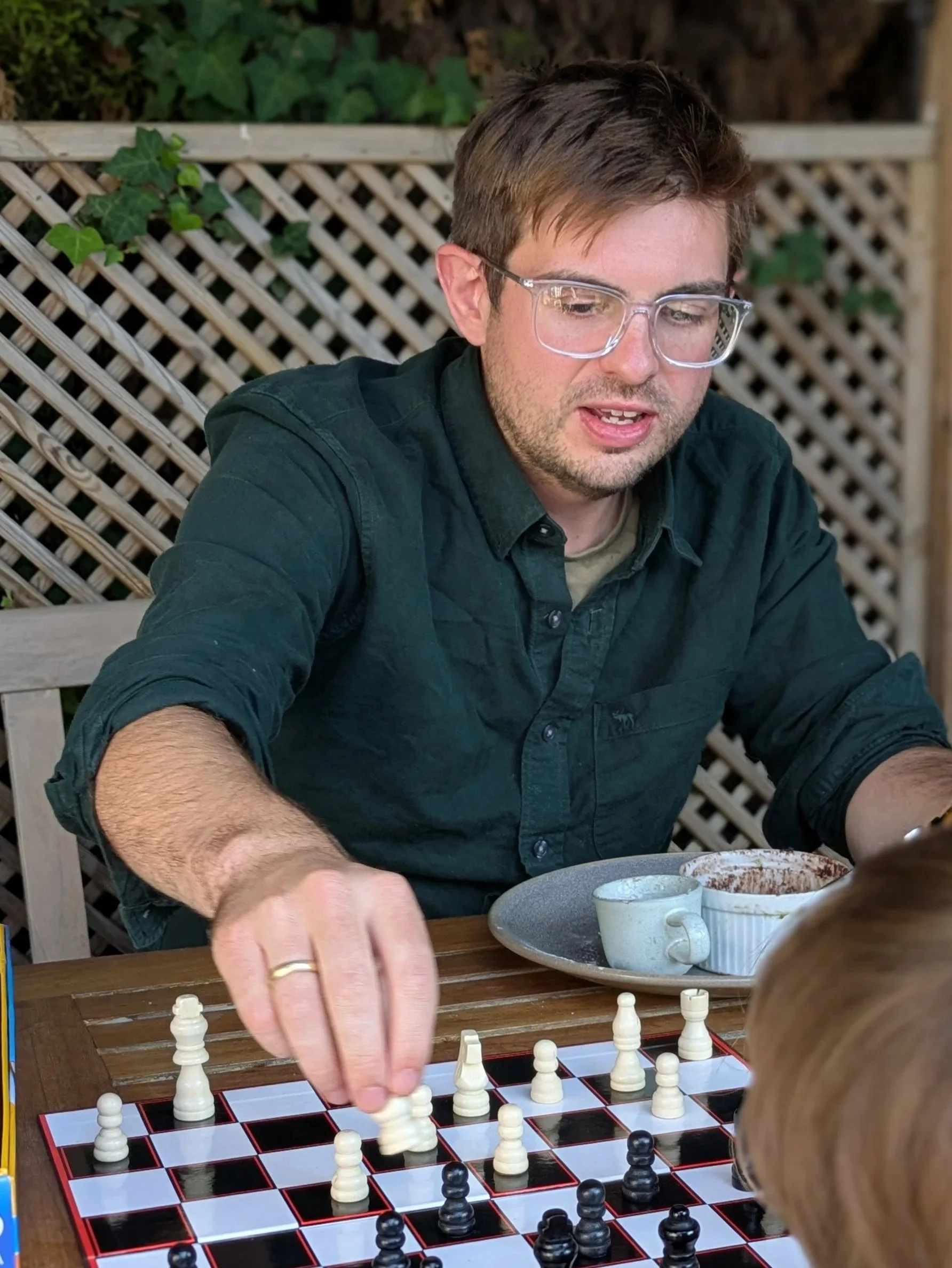 A man with glasses and a beard playing chess outdoors, with a child observing, on a checkered chessboard