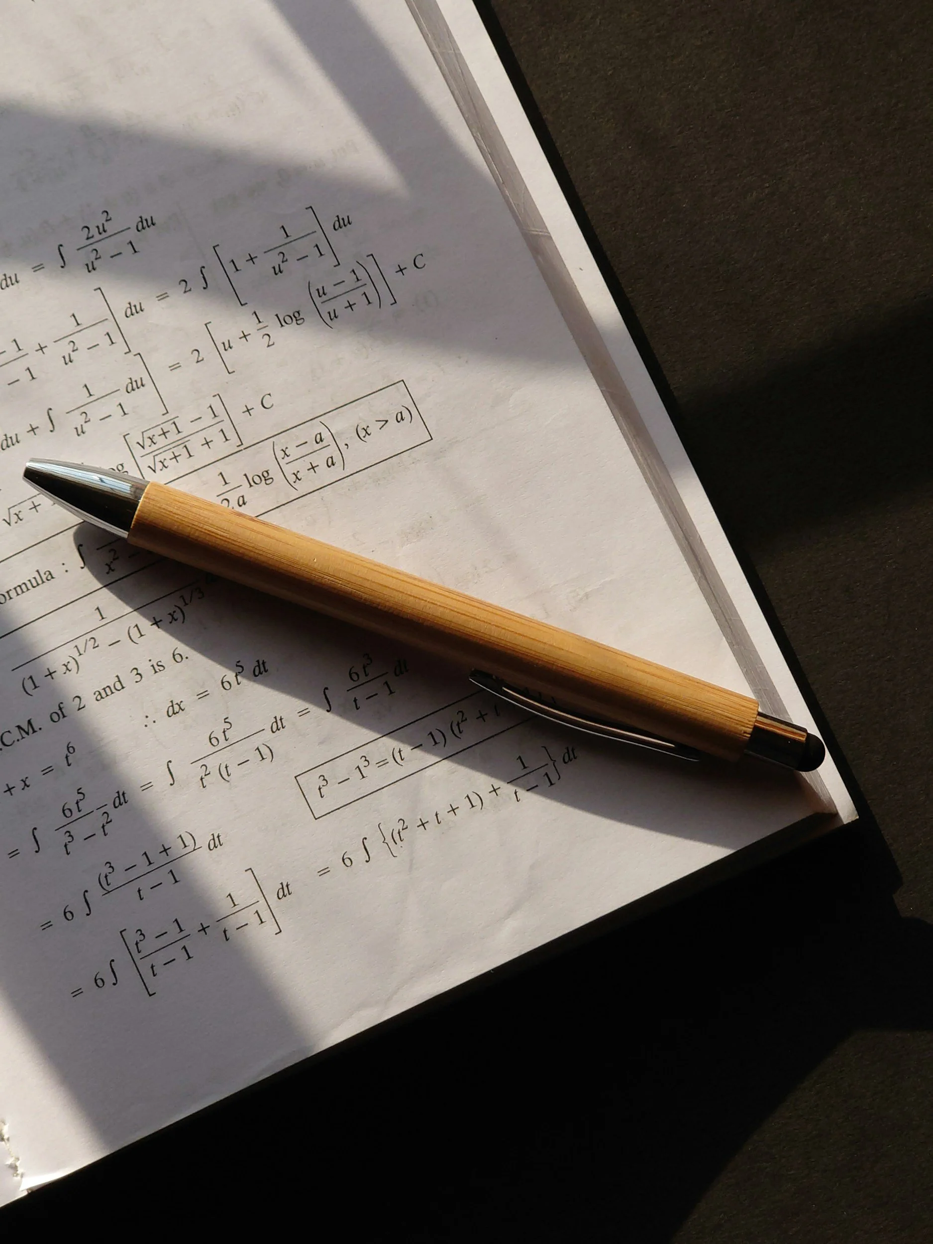 A page of mathematical equations and formulas, with a wooden pen resting on the page, and a shadow cast over part of the page.