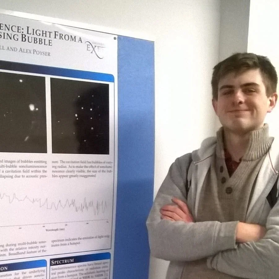 Young man with crossed arms standing next to a science poster about light from a bubbling experiment.