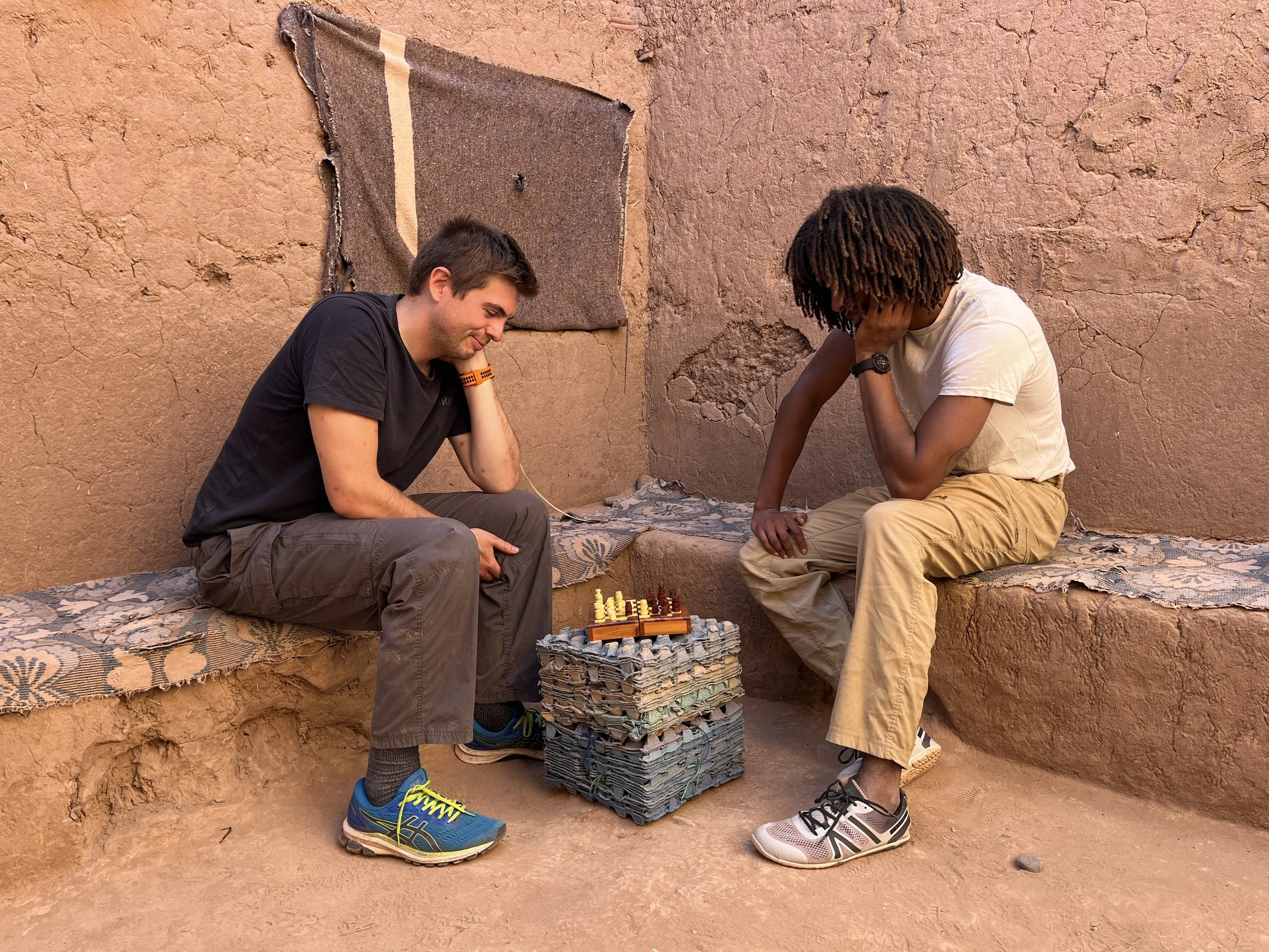Two young men sitting against a sandy, adobe wall playing chess on a small wooden chessboard placed on top of a stack of cardboard boxes in the desert.