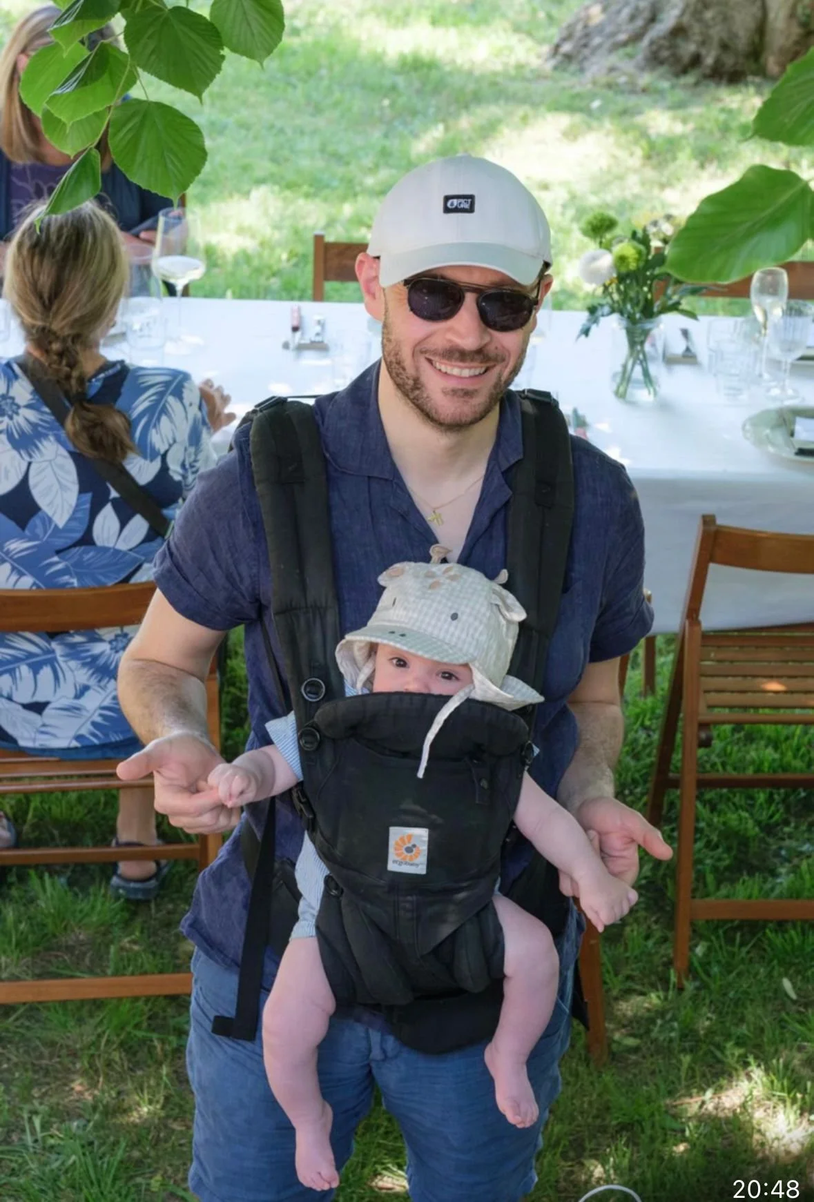 A man wearing sunglasses, a baseball cap, and a backpack is holding a baby in a carrier. The baby is wearing a hat with animal ears and is looking at the camera. They are outdoors near a table set for a meal with wine glasses and a bouquet of flowers.