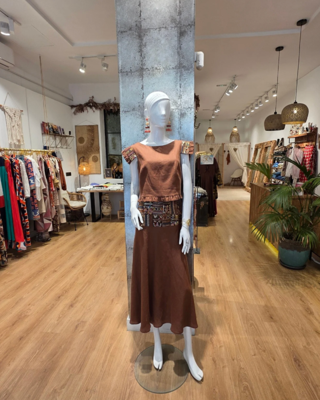 Maniquí con vestido largo y top de color marrón en una tienda de ropa con suelo de madera y decoraciones en tonos naturales.