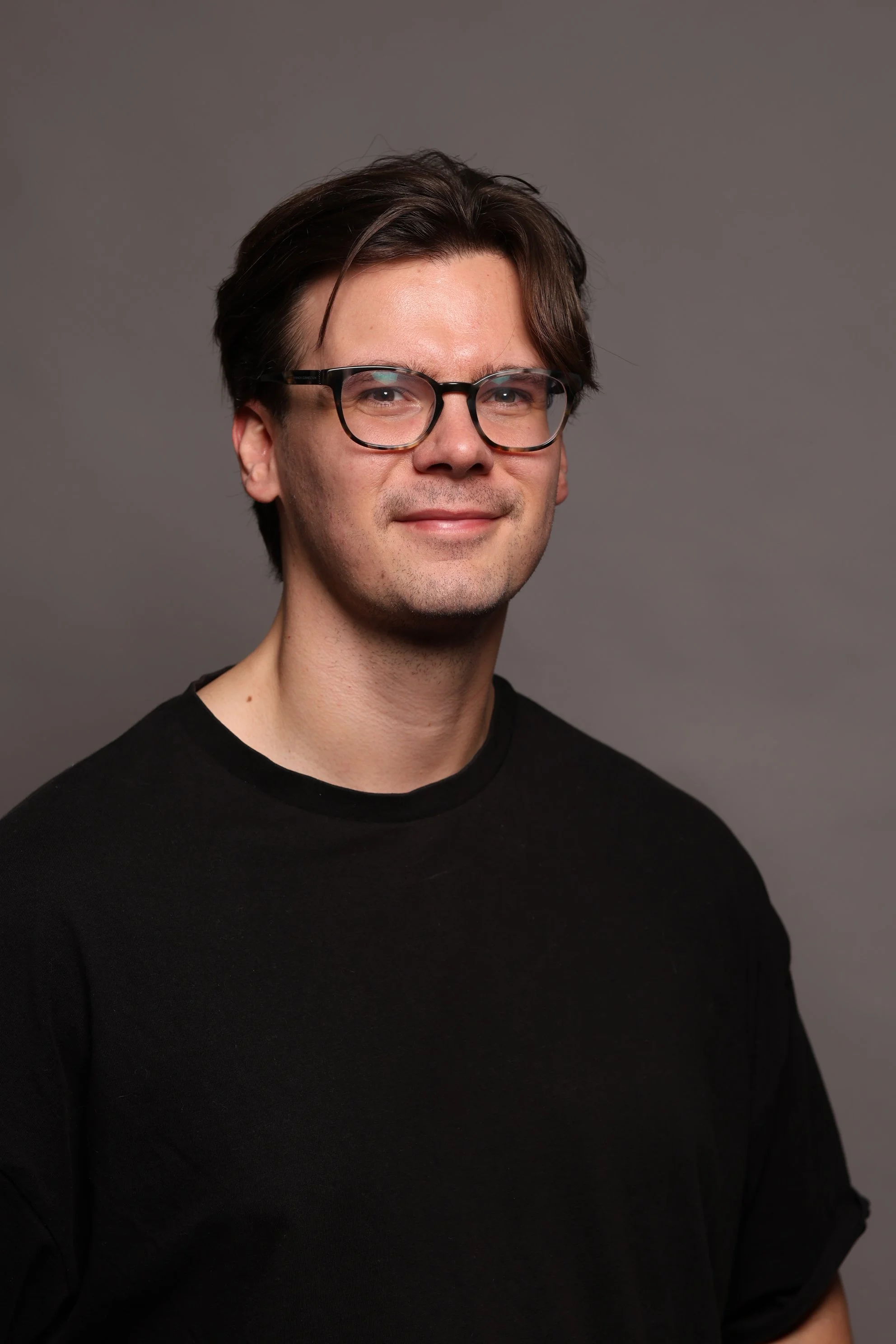 Portrait of a young man with brown hair wearing glasses and a black shirt, smiling slightly, against a plain gray background.