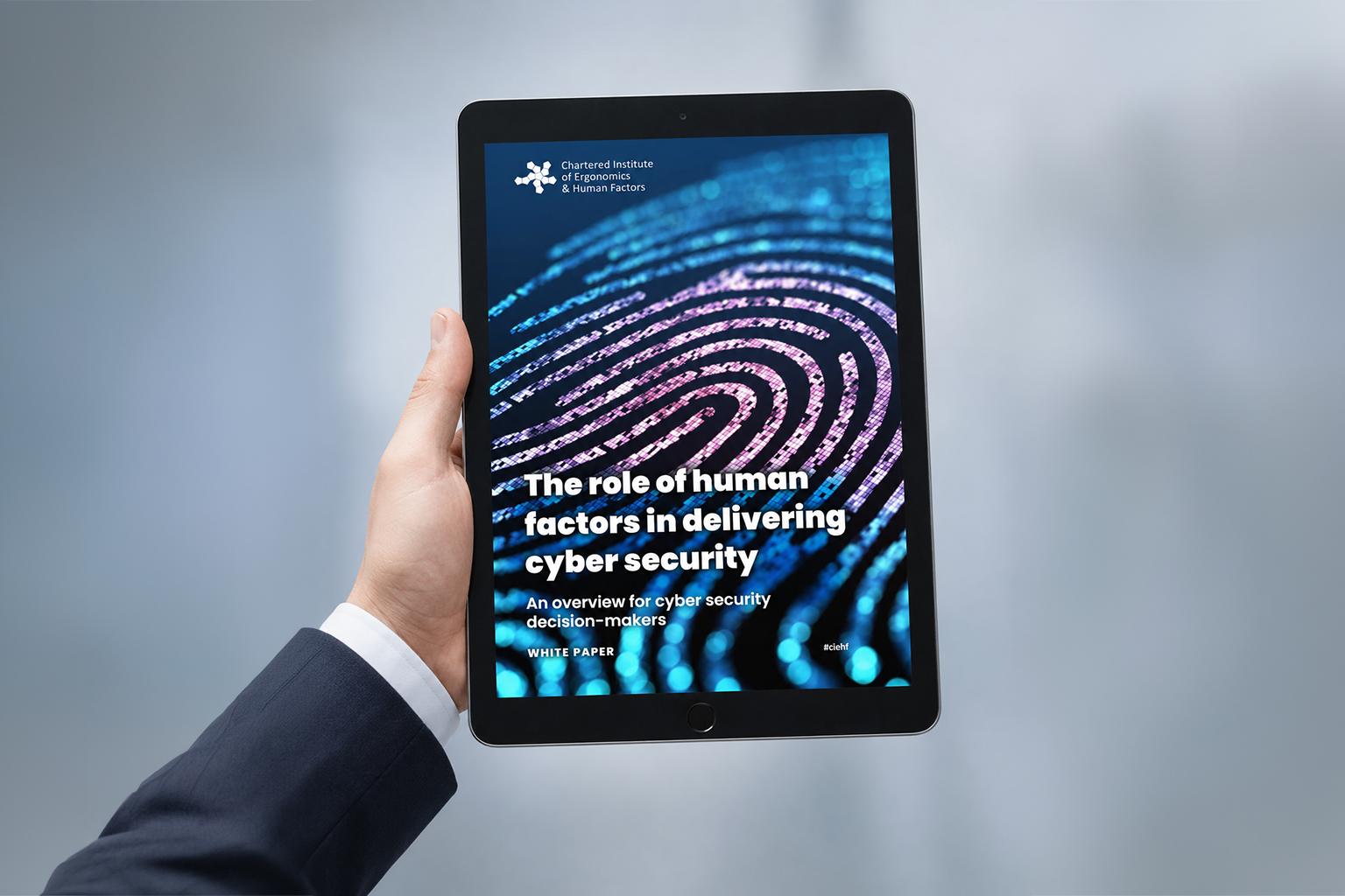 Hand holding a tablet displaying a cover page titled 'The role of human factors in delivering cyber security' from a white paper by the Chartered Institute of Ergonomics & Human Factors, featuring a digital fingerprint graphic.