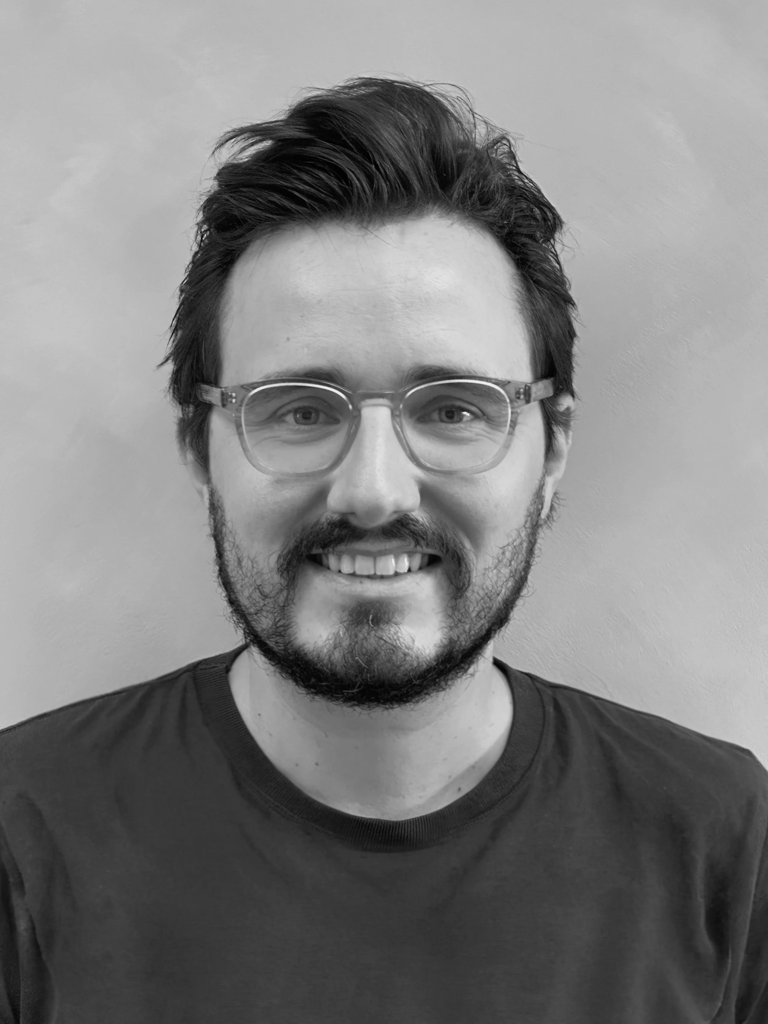 Black and white photo of a man with dark, styled hair, glasses, a beard, and a mustache. He is smiling, showing his teeth, and wearing a dark shirt.