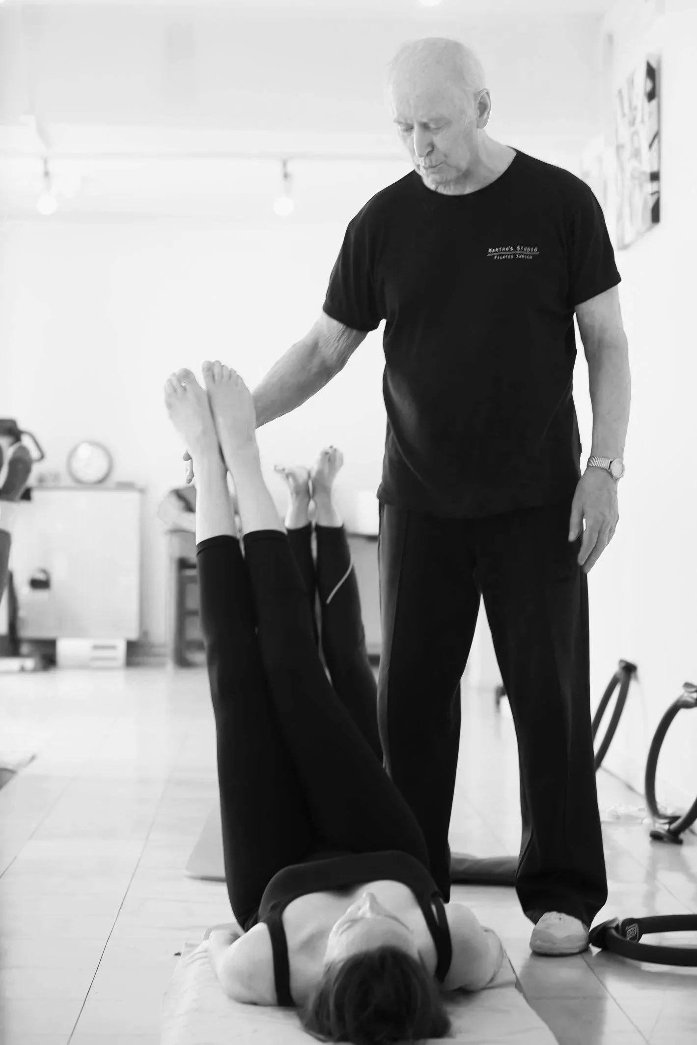 Un homme avec un t-shirt noir enseigne une séance de physiothérapie à une femme allongée sur un tapis, dans une salle d'entraînement. L'homme observe attentivement la femme qui réalise un exercice sur le dos, en utilisant ses jambes comme support.