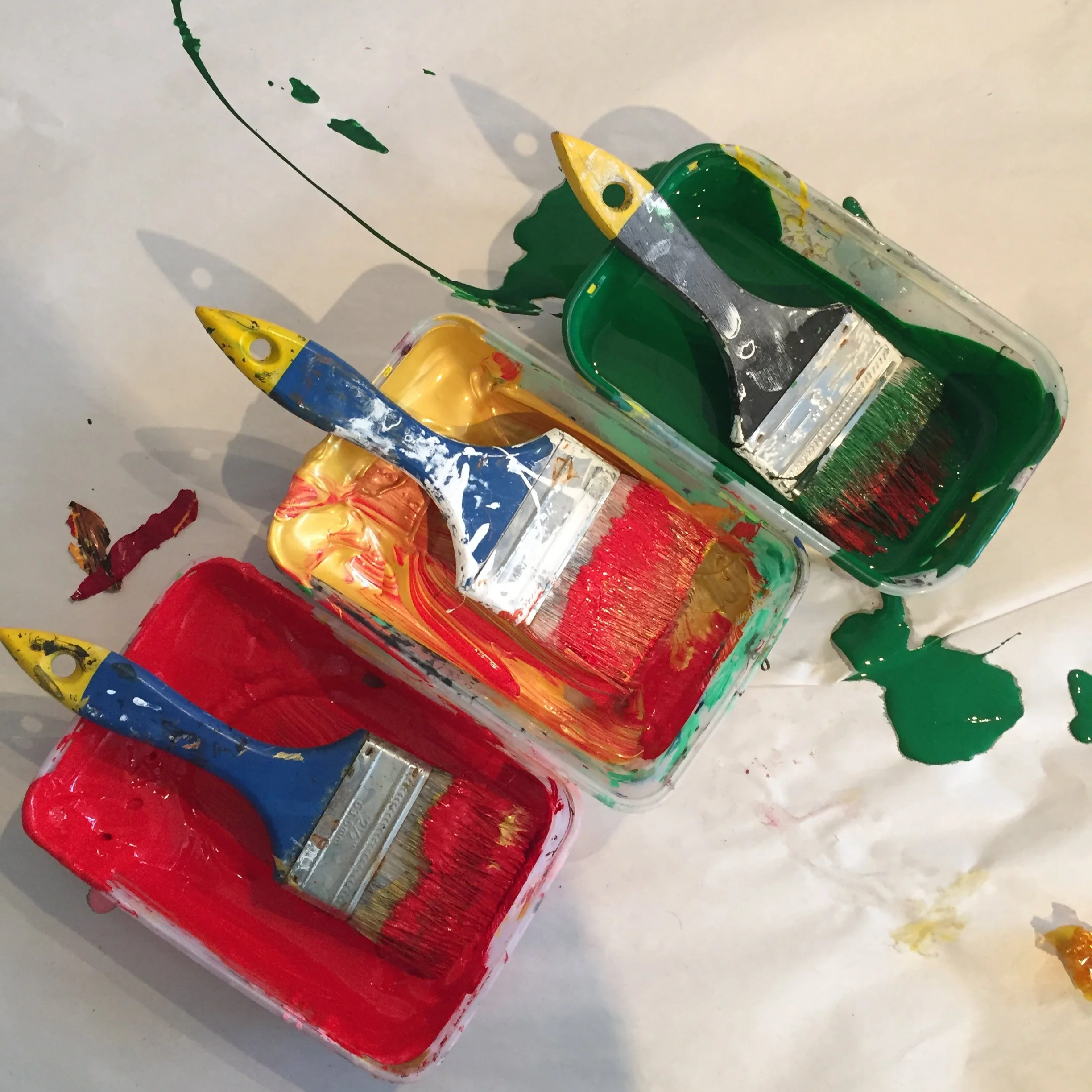 Open paint cans filled with red, green, yellow, and blue paint, with paintbrushes and spilled paint on a white surface.